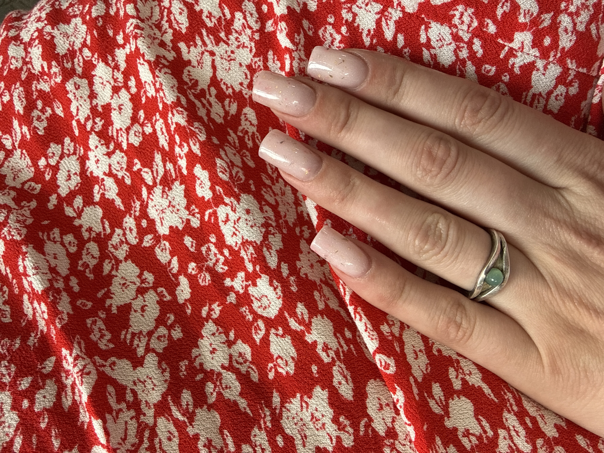 A person's hand, adorned with a silver ring featuring a green stone and light pink, square-shaped artificial nails with gold flecks, rests on a red fabric with a white floral pattern. The fabric is wrinkled and appears to be a garment or upholstery. The lighting suggests an indoor environment, possibly a home, given the casual presentation. The background is solely the patterned fabric, with no other elements of the environment visible. The image is a close-up, focusing on the hand and the texture of the fabric.
