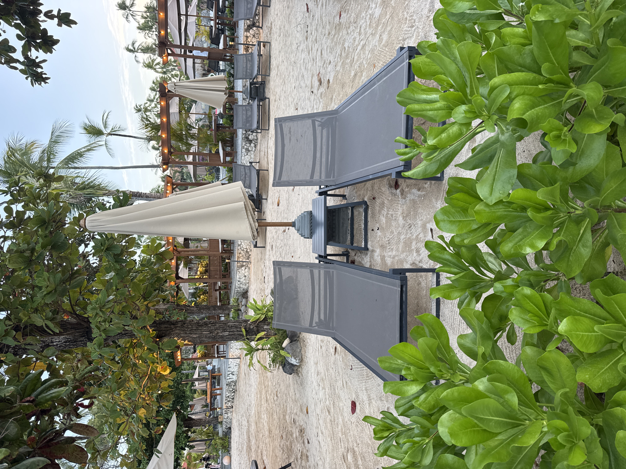 The image depicts a tranquil beach scene at sunset in Kuta Selatan, Indonesia. In the foreground, lush green foliage partially obscures two dark grey lounge chairs positioned on the sandy beach. A closed, beige umbrella stands between the chairs, accompanied by a small, dark grey side table.
In the mid-ground, more lounge chairs and closed umbrellas are visible, suggesting a resort or hotel setting. Beyond these, several wooden pergolas with thatched roofs and string lights create a warm ambiance. Palm trees are scattered throughout the scene, with some in the background showcasing the vibrant tropical environment. The sky is a soft blue, indicative of the evening hour.
The scene is devoid of any people actively engaged in activities, contributing to the overall sense of serenity. The lighting suggests the golden hour, casting a gentle glow on the beach and surrounding elements. The sand appears clean and well-maintained, and the vegetation is healthy and vibrant.