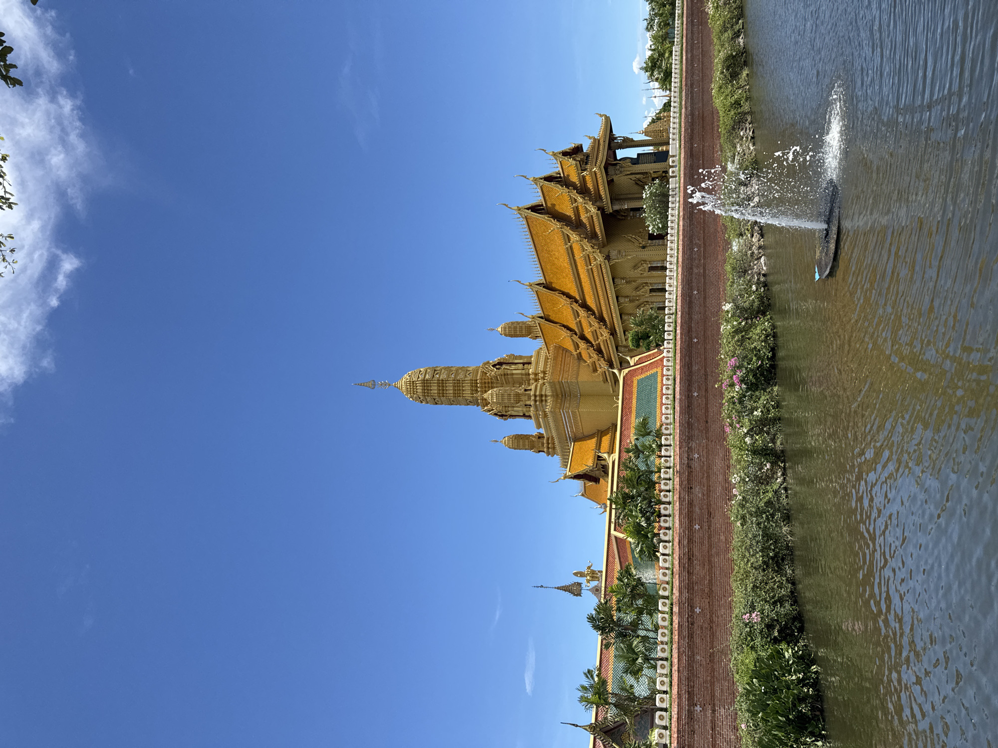 The image captures the golden pagoda and buildings of Wat Asokaram, a Buddhist temple in Bang Pu Subdistrict Municipality, Thailand. The temple complex features elaborate golden architecture with tiered roofs and ornate detailing. A body of water, possibly a pond, runs alongside the temple grounds, with a fountain spraying water into the air. A brick wall topped with greenery and flowers separates the temple complex from the water. The sky is a clear blue with a few wispy clouds.