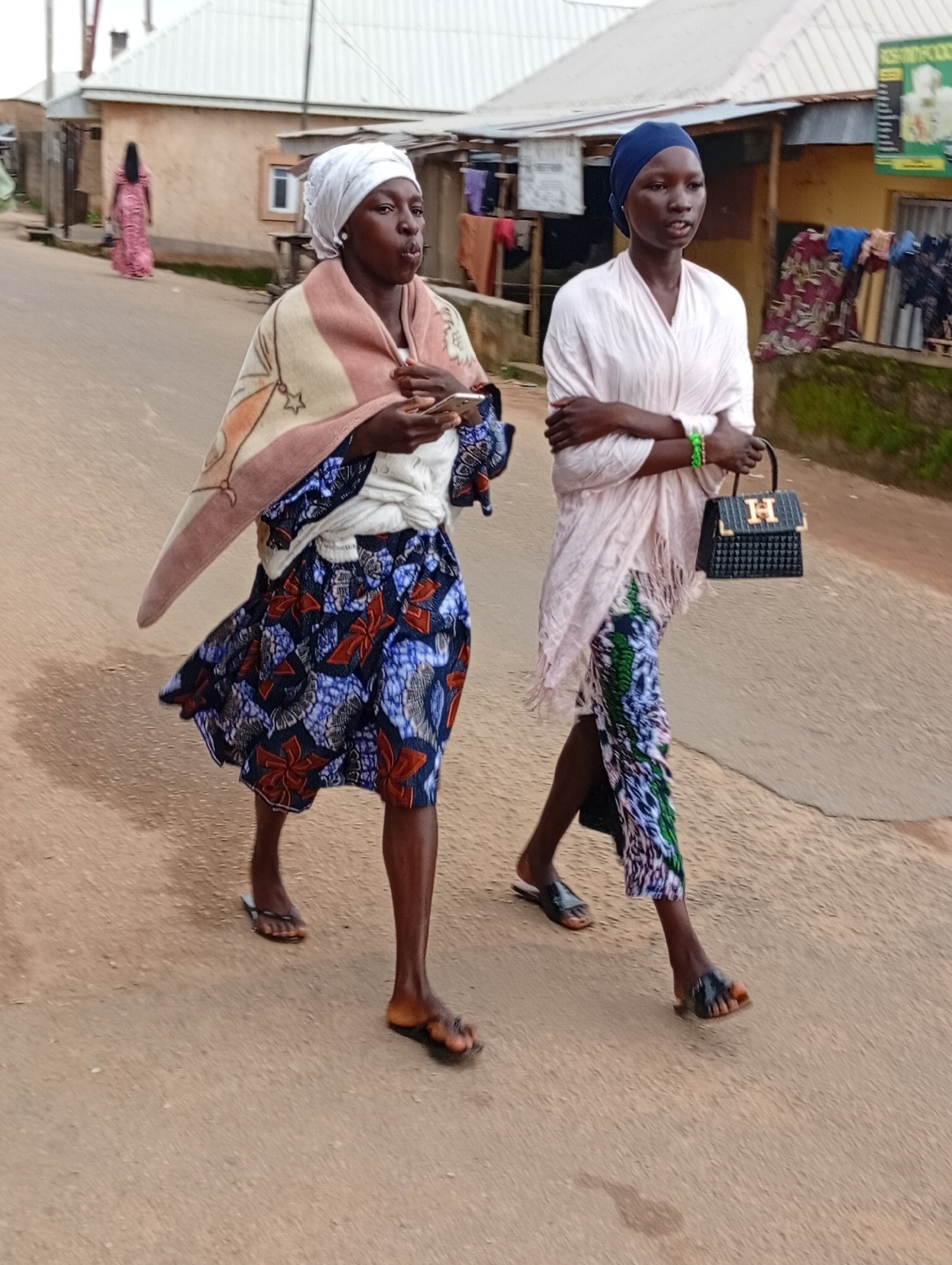 In Jos, Nigeria, two young women walk side-by-side on a dusty, paved road. The woman on the left wears a white headscarf, a blanket draped over her shoulders, and a vibrant blue and red patterned skirt. She holds a mobile phone in her right hand. The woman on the right wears a navy blue headscarf and a light pink shawl over a long-sleeved shirt. Her patterned skirt features green, blue, and white designs. She carries a black handbag with an "H" emblem on it and wears a green bracelet. The background shows buildings with corrugated metal roofs, some with laundry hanging outside. The environment appears to be a residential area with a muted color palette of earth tones and building materials.