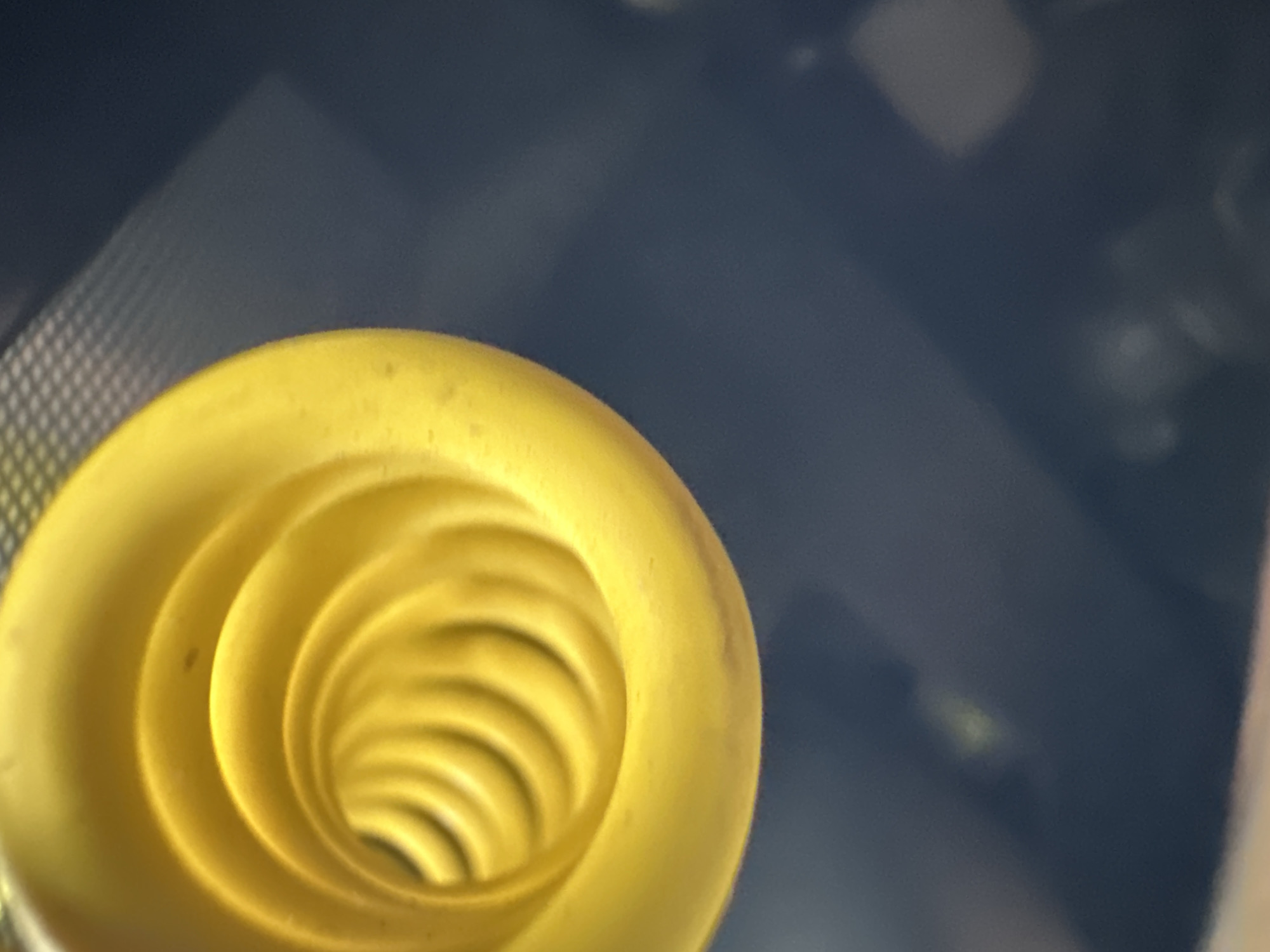 The image shows a close-up, abstract view of a bright yellow, coiled object against a dark, out-of-focus background. The object appears to be a funnel or a spiral tube, with concentric ridges creating a sense of depth. The lighting highlights the smooth, curved surfaces of the yellow object, giving it a glossy appearance. The background is a dark, indistinct expanse, with some blurry lighter areas that could be reflections or distant objects. There are no people or clear signs of a specific location. The mention of Shatava, Ukraine, is not supported by any visual evidence in the image itself.