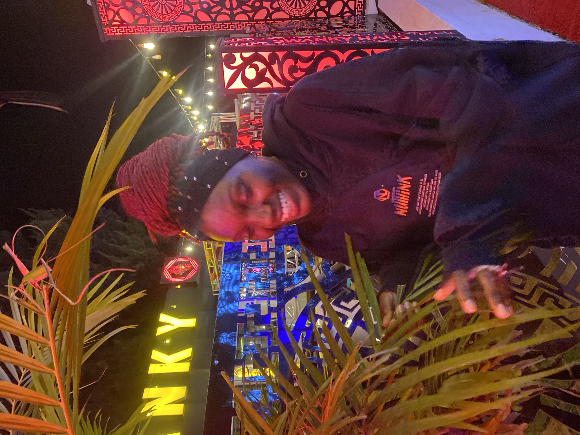 A young Black woman with dark skin, wearing a black hoodie and a black headband with embellishments, stands in front of a palm frond, smiling with her mouth open and teeth showing. Her hair is braided in a deep red color and styled in a bun on top of her head. The background features neon signs and decorative elements, suggesting an outdoor nighttime setting. A bright yellow sign that reads "INKY" is visible in the upper left, and a red, geometric light fixture is on the right. Strings of yellow lights are strung across the background. The woman's right hand is visible, reaching out towards the palm frond, with her fingers slightly spread. The overall lighting is dim, with the neon signs and light fixtures providing the primary illumination.