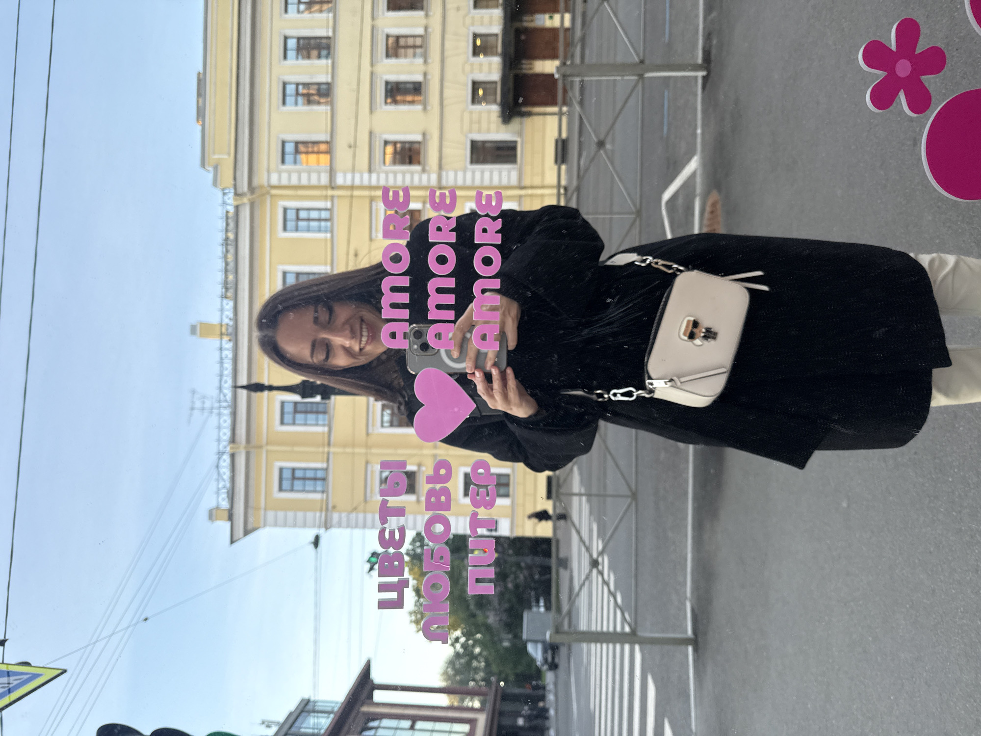 A woman is reflected in a shop window in Saint Petersburg, Russia, taking a selfie with her phone. She is wearing a black coat and cream-colored pants, and a small cream-colored crossbody bag is visible. Pink text on the window reads "ЦВЕТЫ ЛЮБОВЬ ПИТЕР" and "AMORE AMORE AMORE." A pink heart sticker is also on the window. Through the window, a yellow building with arched windows and balconies is visible across a street with a pedestrian crossing and a traffic light. The sky is overcast.