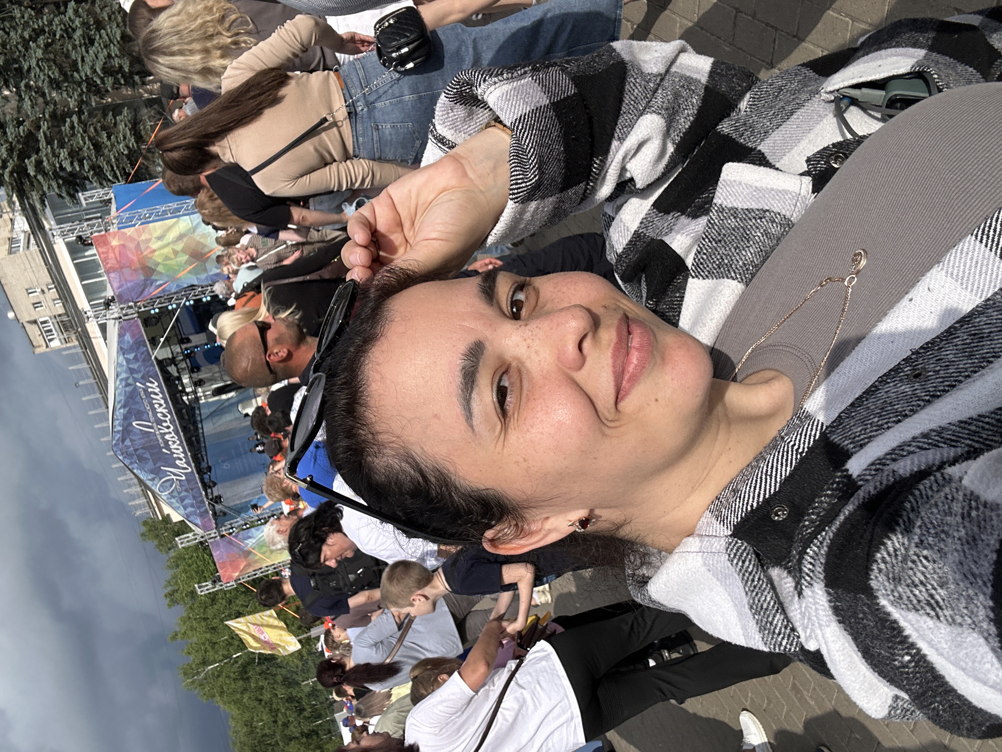 A woman with dark hair smiles at the camera in a selfie taken outdoors. She is wearing a grey t-shirt and a black and white plaid jacket. She is holding her hand up to her hair. Behind her, a crowd of people are gathered in front of a stage with a banner that reads "Чайковский". The sky is cloudy.