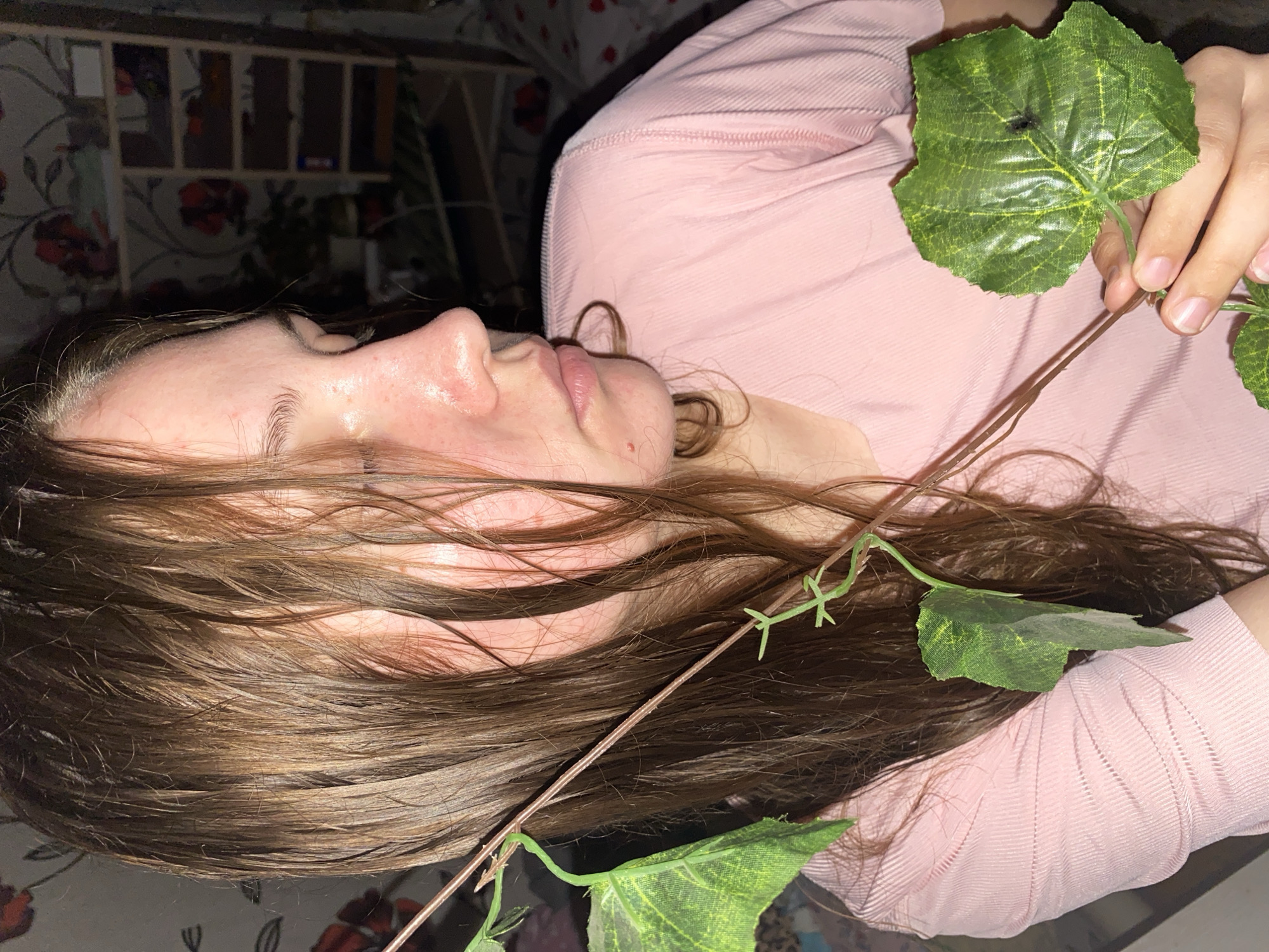 A young woman with wet, brown hair is shown in a close-up, profile view. She is wearing a pink, ribbed shirt. A decorative vine with green leaves is held in her right hand, draped across her chest and hair. In the background, a shelf unit with various items is visible, along with wallpaper with a floral pattern. The scene appears to be indoors. No specific location in Вассята, Russia can be determined from the visual information.