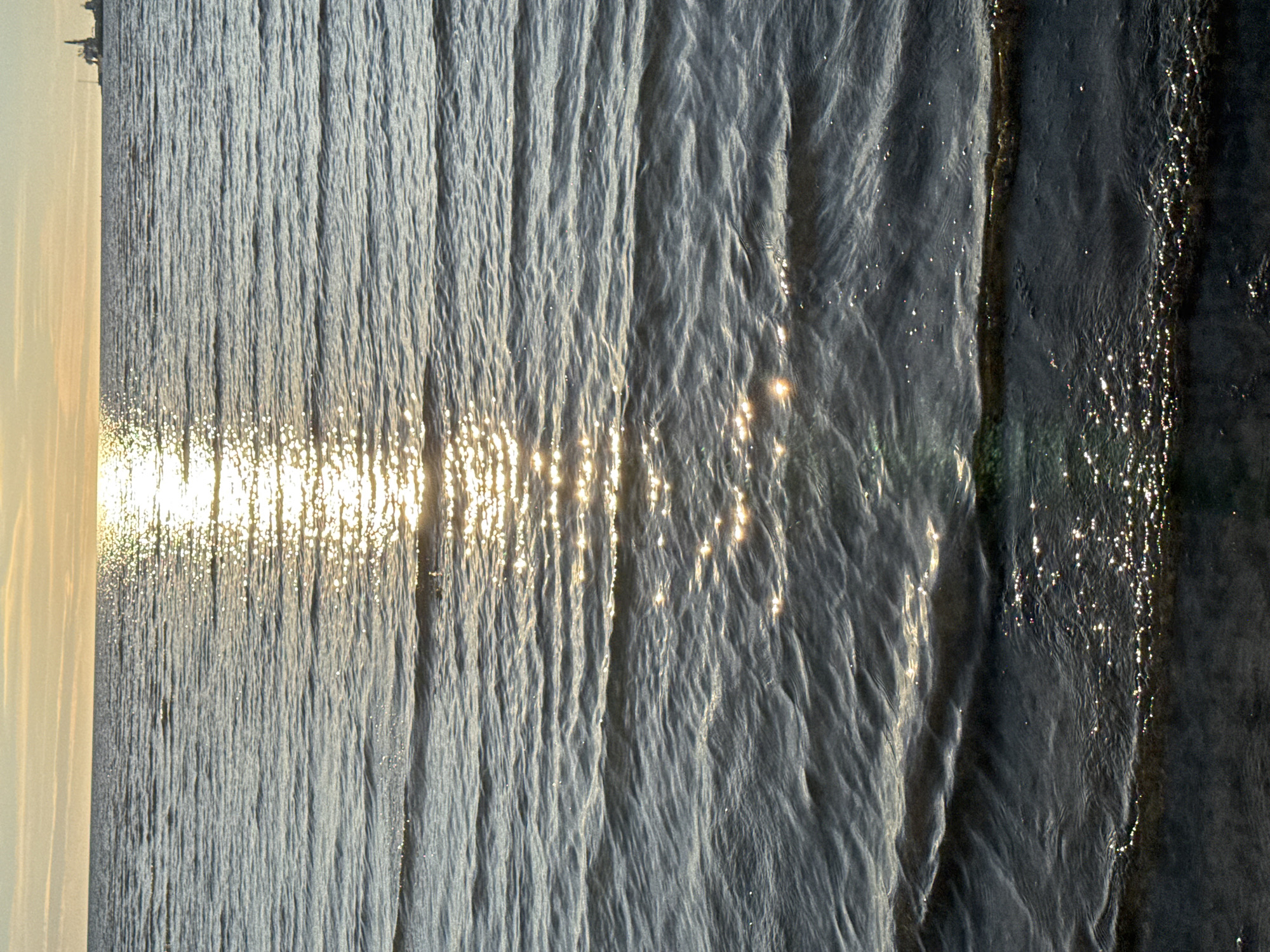 The image depicts a wide expanse of water, likely a sea or large lake, under a pale, hazy sky. The sun, low on the horizon and partially obscured by clouds, casts a bright, shimmering reflection across the surface of the water. The water itself is characterized by numerous small, rhythmic ripples, creating a textured and dynamic visual. In the far distance, along the horizon, a faint silhouette of what appears to be industrial structures or ships is visible. The foreground shows the edge of the water meeting a dark, indistinct shore. The scene evokes a sense of calm and the vastness of nature, with a hint of human activity present in the distant structures. This scenery is consistent with the coastal areas of Tallinn, Estonia, particularly views looking out towards the Baltic Sea.