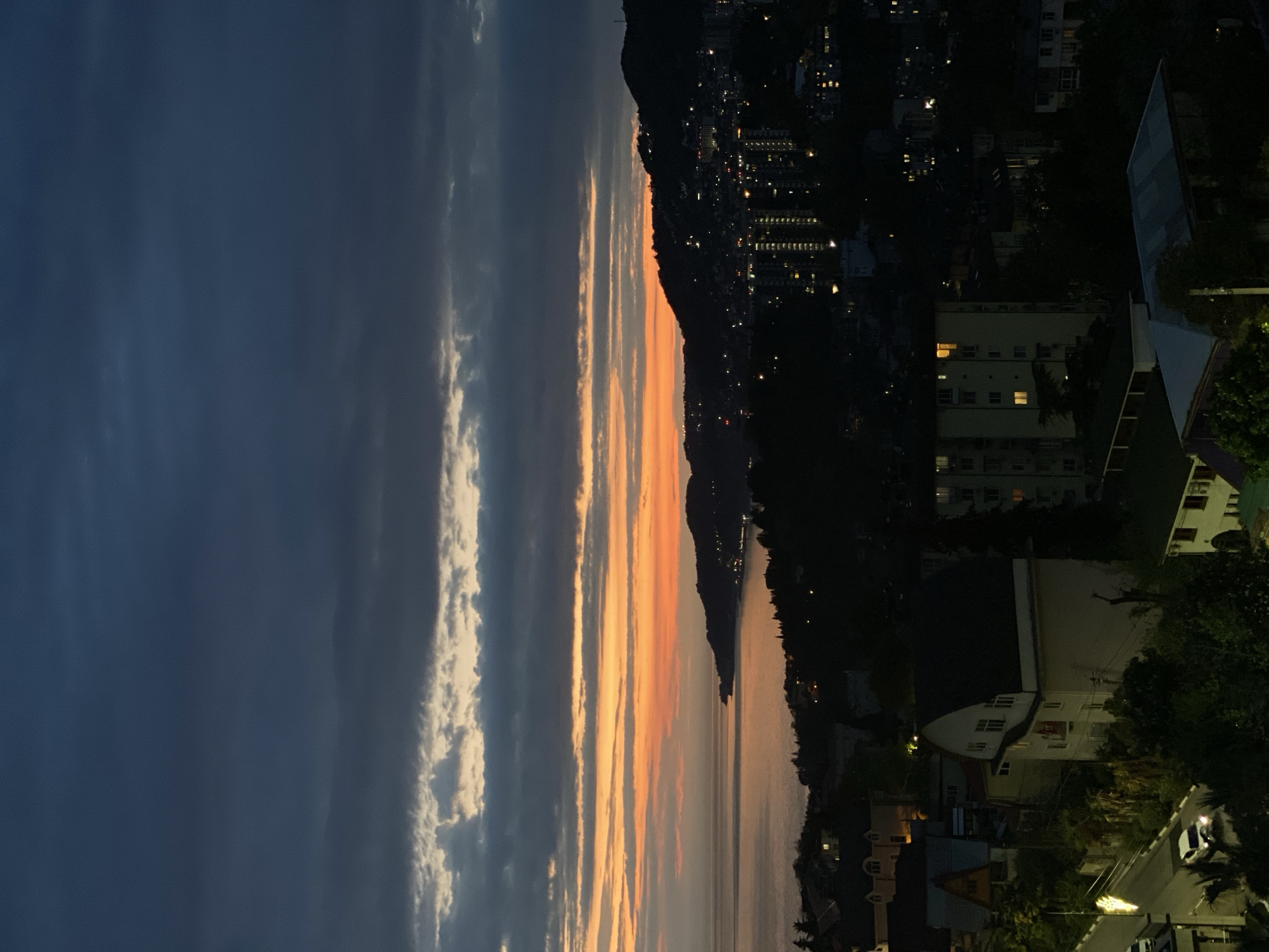 The image captures a cityscape during a twilight or sunset. The sky on the left side is predominantly dark blue with layers of light grey clouds, transitioning into orange and yellow hues towards the center, suggesting the sun setting or having set. The right side of the image features a dark, mountainous landscape dotted with the lights of buildings. Lower down, buildings and foliage are vaguely discernable in the darkness, with hints of streetlights and a passing car. The scene likely depicts a view over a body of water or valley towards a city area built on the slopes of a hill or mountain range. While the image hints at urban development amidst natural landscapes, the absence of significant detail makes pinpointing the location as Sochi, Russia speculative.