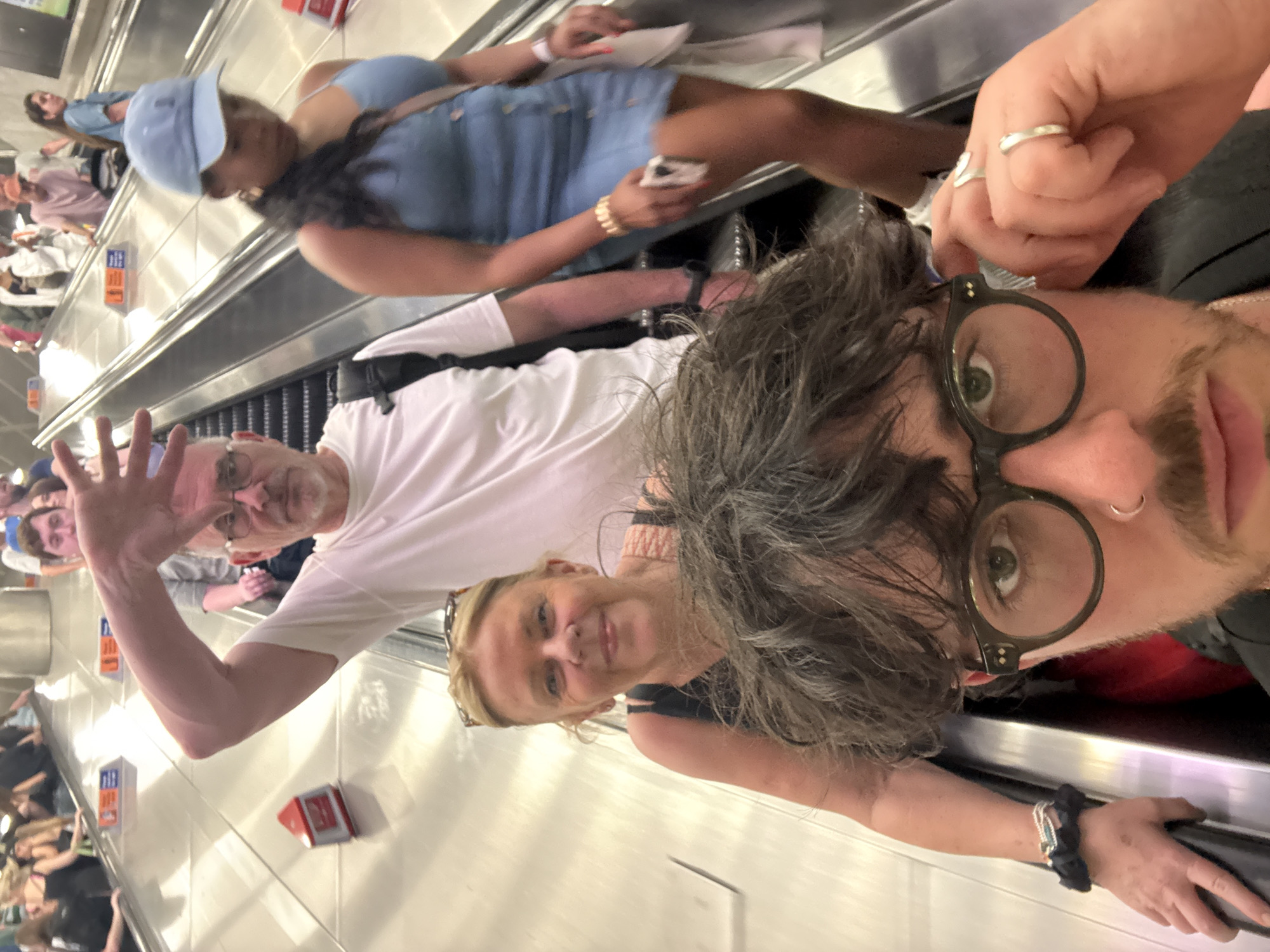 A group of people are traveling on an escalator. One man with glasses and a white shirt is waving at the camera. A woman with blond hair is standing beside him. In the foreground, a person with glasses and dark hair is taking a selfie. Behind them, another woman in a blue dress and hat is using a cell phone. The escalator is located in an area with red and blue signs on the side.