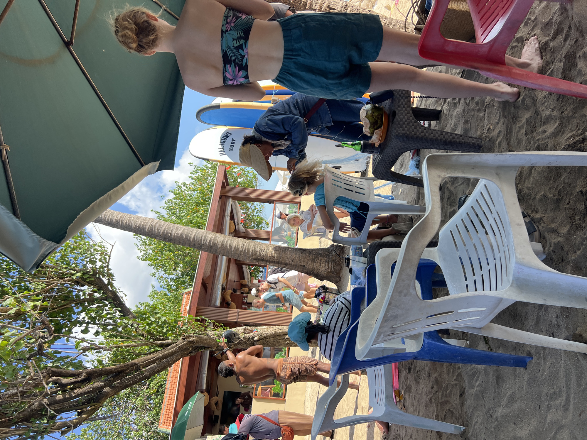 This is an outdoor scene on a sandy beach in Kuta, Indonesia. Several people are present, including a woman getting her hair styled by another woman. A man is standing shirtless, and other individuals are seated at tables or walking in the background. Surfboards are propped up near a building that appears to be a cafe or shop. Beach chairs are scattered on the sand. The atmosphere suggests a casual beachside gathering or activity.