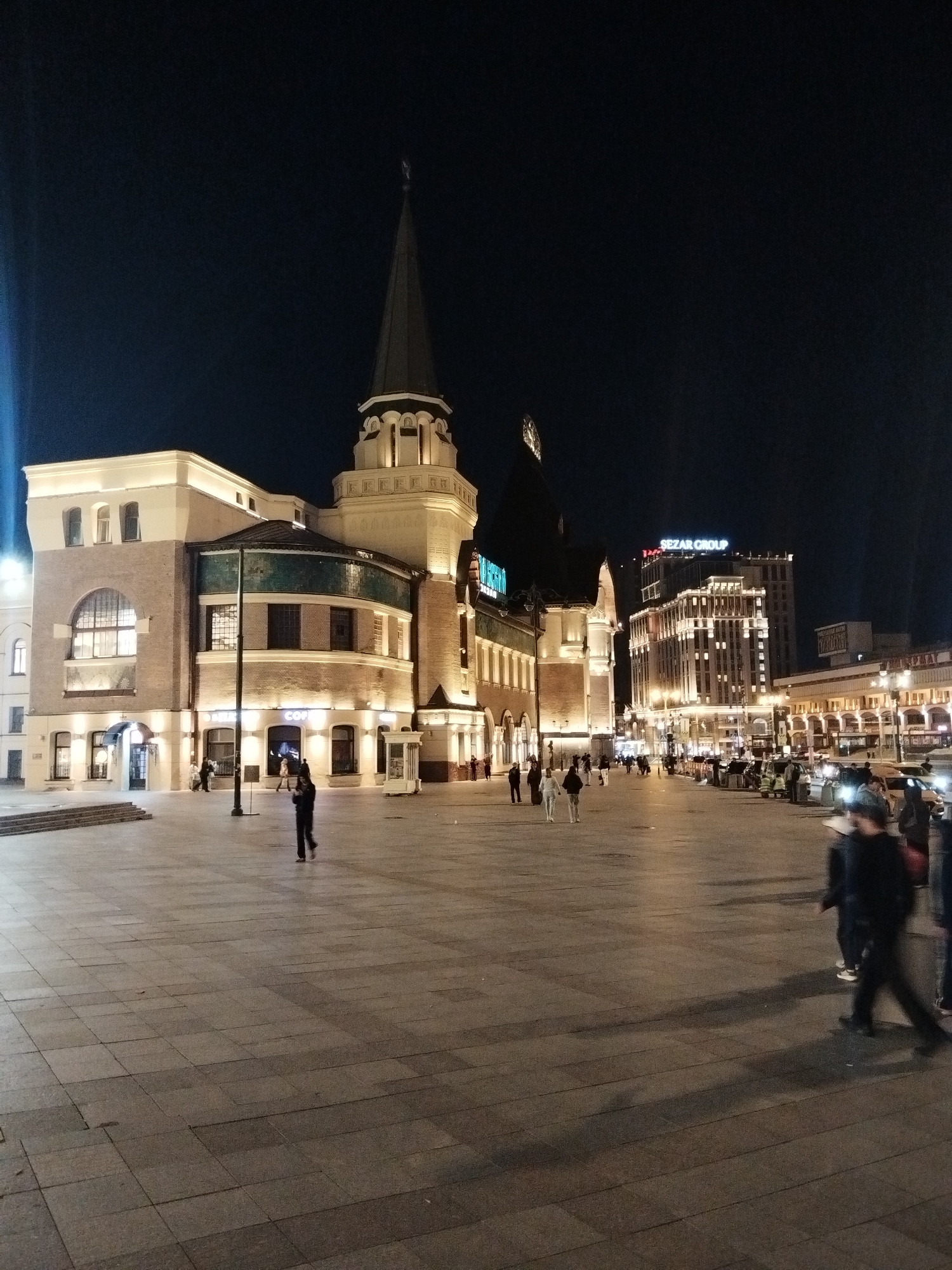 The image depicts a night scene in Moscow, Russia, specifically at Komsomolskaya Square, also known as "Three Station Square." In the foreground, a large, paved plaza is visible, with several people walking across it. The ground is illuminated by artificial lighting, casting long shadows.
Dominating the left side of the frame is a building with distinctive architecture, featuring a tall, spire-like tower. This building appears to be a historical structure, possibly part of the Leningradsky Railway Station, given its proximity to the square. The facade is illuminated with warm yellow lights.
In the background, to the right, stands a modern, multi-story building with a prominent sign that reads "SEZAR GROUP." This building is also brightly lit, showcasing its windows and architectural details. Further to the right, another building with arched windows and illuminated columns lines the street.
The overall atmosphere is that of a busy urban space at night, with a blend of historical and modern architecture, illuminated by various light sources. The presence of people suggests ongoing activity in the area.