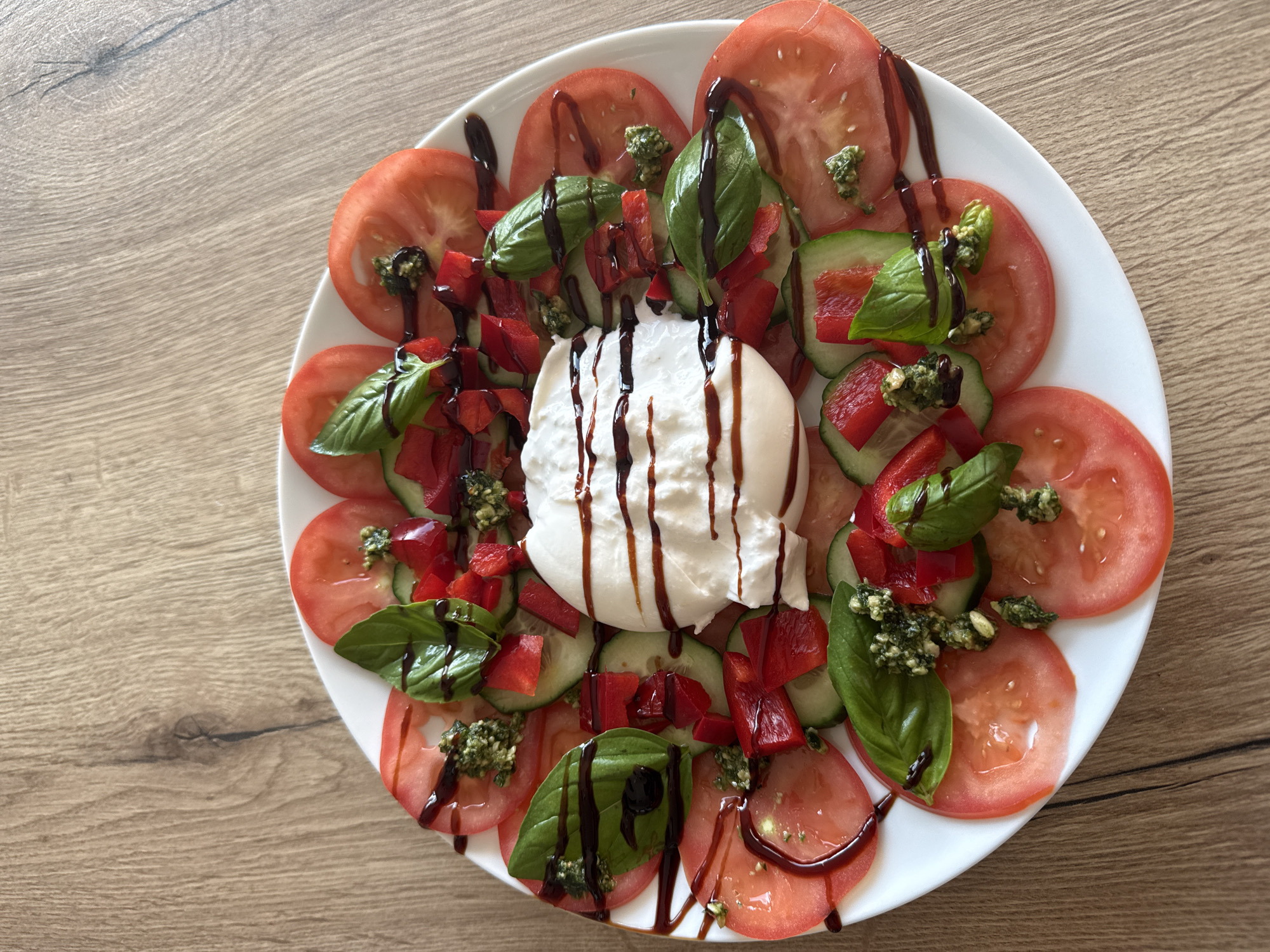 A top-down view of a white plate filled with a colorful salad on a wooden surface. The salad features sliced tomatoes arranged in a circular pattern around the edge of the plate. Inside the ring of tomatoes, there are cucumber slices, diced red bell peppers, and fresh basil leaves. Small portions of pesto are scattered throughout the salad. At the center of the plate is a white, spherical mass, possibly burrata cheese, drizzled with a dark balsamic glaze, which is also drizzled across the rest of the salad. The scene is illuminated with natural light, creating shadows that suggest the photo was taken indoors near a window. The wooden surface has a light brown, linear grain pattern. The location is Pétange, Luxembourg.