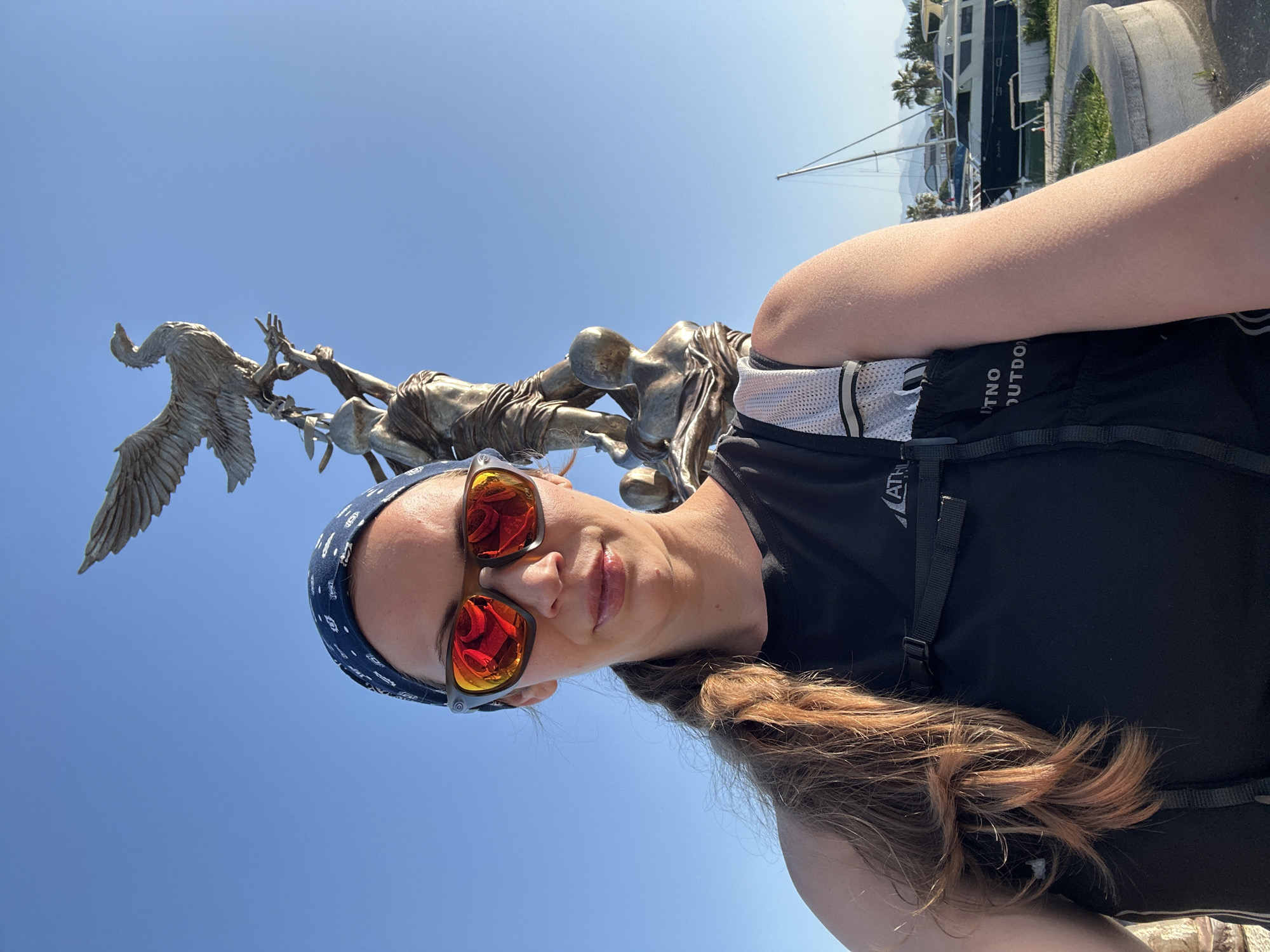 The image captures a woman with long, auburn hair styled in a low braid, wearing a black sleeveless athletic shirt and a black backpack. She sports a blue patterned bandana and reflective sunglasses with orange lenses. She is positioned in front of a bronze sculpture depicting figures and a bird, set against a clear blue sky. In the background, a marina with docked yachts and greenery can be observed. The location is Fethiye, Turkey, evidenced by the marina and architectural style.