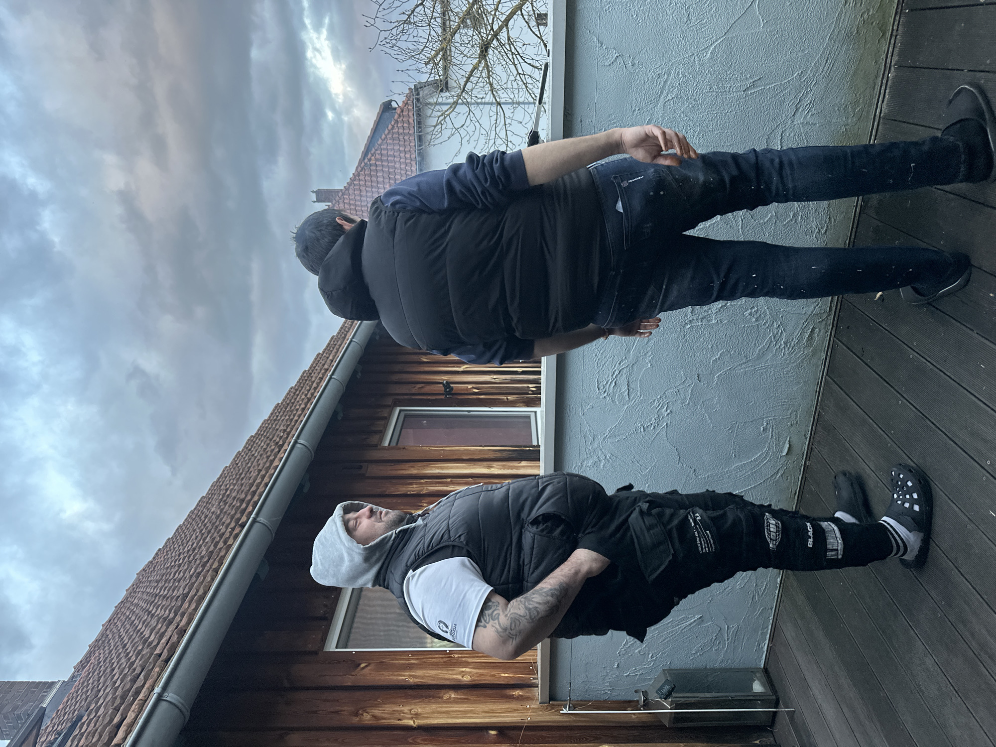 Two men stand on a wooden deck or patio in Heinebach, Germany. One man wears a white t-shirt, grey hoodie, black vest, black pants with white lettering and designs, black socks, and black Crocs. He has tattoos on his left arm. The other man wears a navy hoodie, black vest, and dark blue jeans with white paint splatters, and black shoes. They stand near a light blue wall and a wooden building with brown tiled roof. The sky is cloudy.