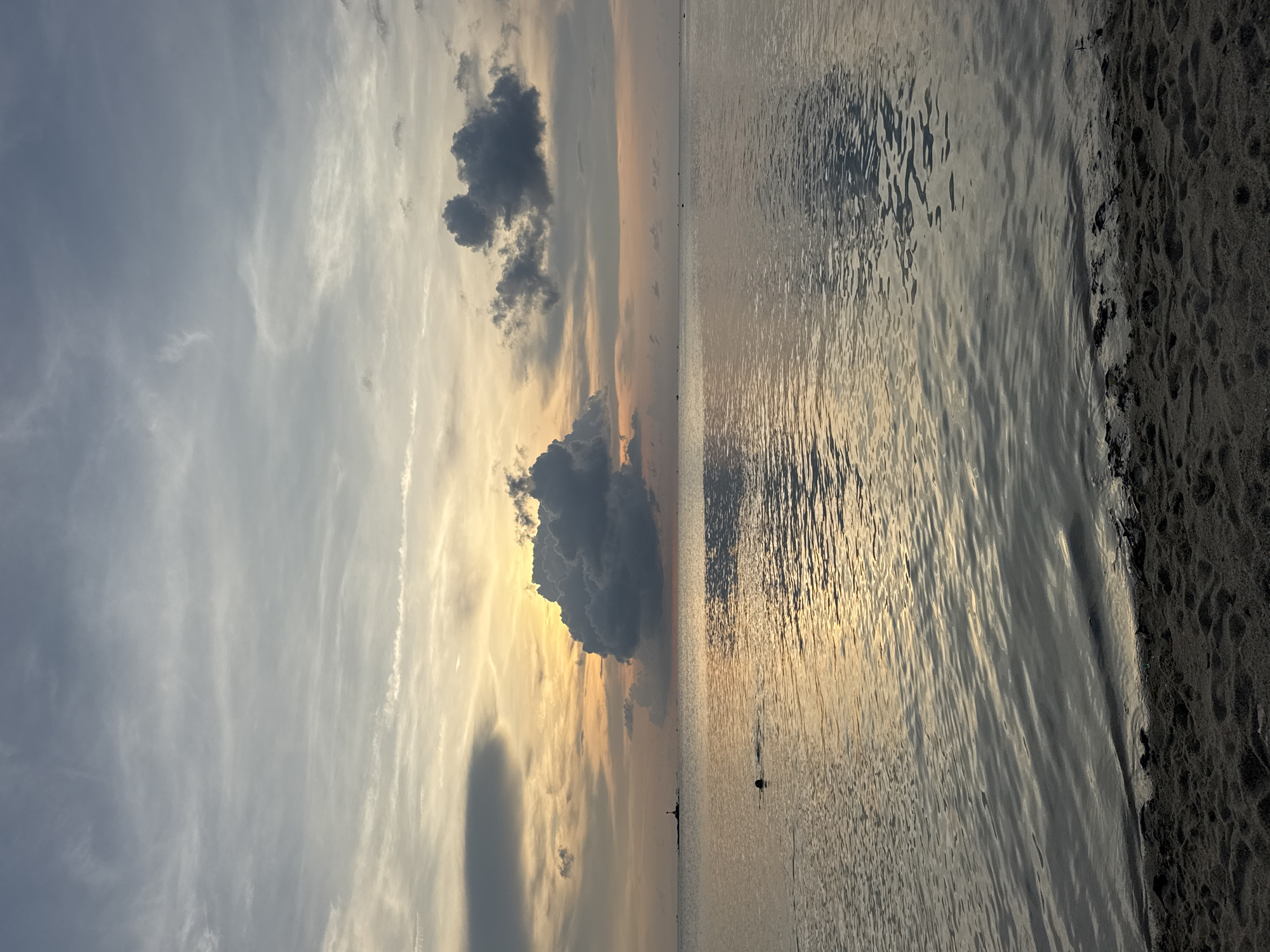 The image depicts a coastal scene near Baan Sri Thanu, Thailand, at what appears to be either sunrise or sunset. The sky exhibits a gradient of colors from gray and white at the top to orange and yellow closer to the horizon line. Several dark, cumulonimbus clouds are visible in the sky.
Below the horizon is the sea, its surface reflecting the colors of the sky. Small waves ripple across the water, creating patterns of light and shadow. A dark object, possibly a buoy or boat, is visible in the water.
The right side of the frame shows a dark, sandy shoreline. The sand is textured, with visible footprints or other impressions.
The image is taken in a vertical orientation.