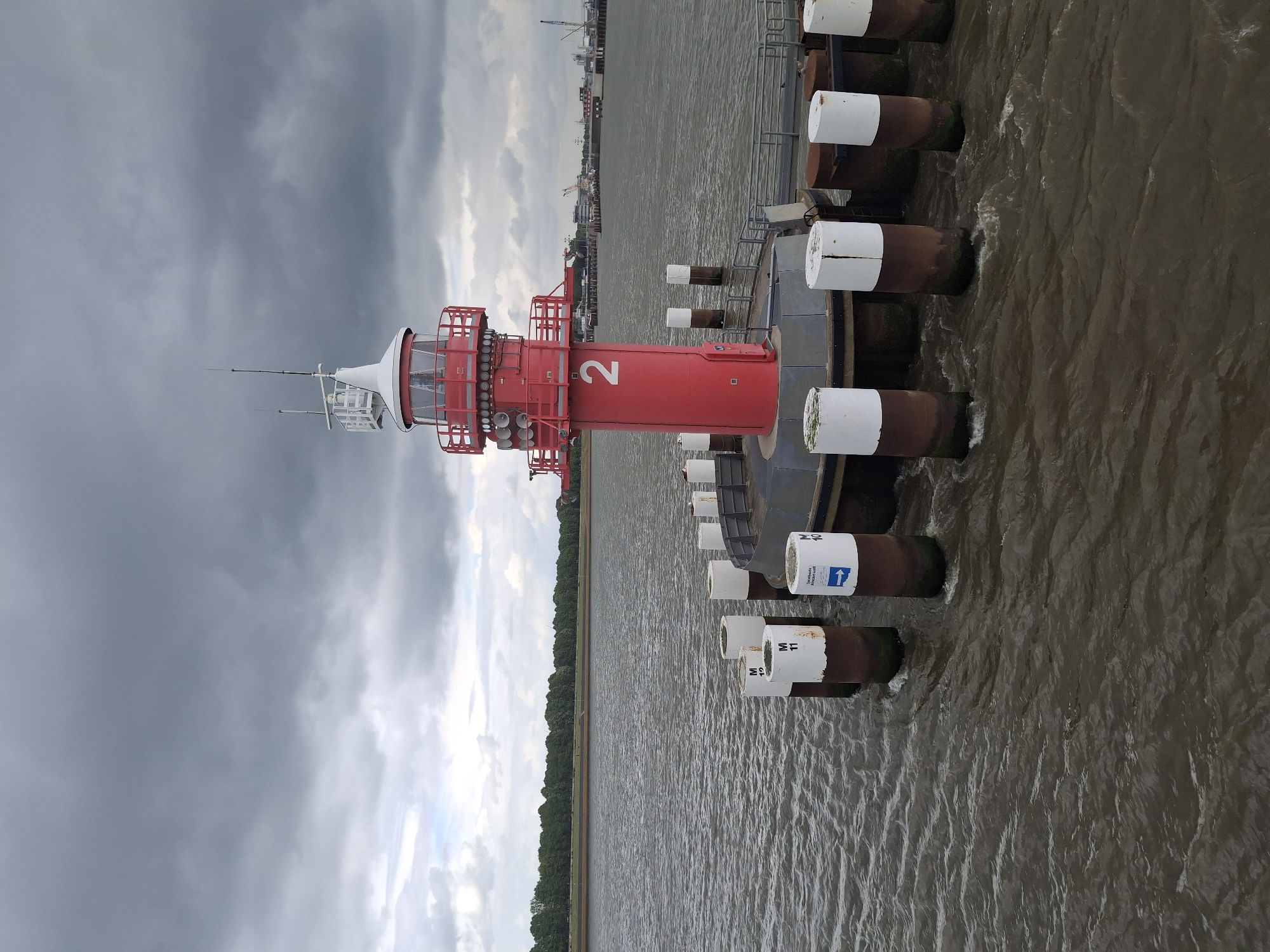The image shows a red lighthouse with the number "2" prominently displayed, situated in the Elbe River near Brunsbüttel, Germany. The lighthouse is mounted on a platform supported by several cylindrical pilings, many with white tops and identifying marks. The river is choppy with a muddy brown color. The sky is overcast with dark, heavy clouds. In the background, the shoreline is visible, with industrial structures and port facilities on the left, and a dense line of trees on the right. The overall impression is of a functional maritime structure in a working port environment on a cloudy day.