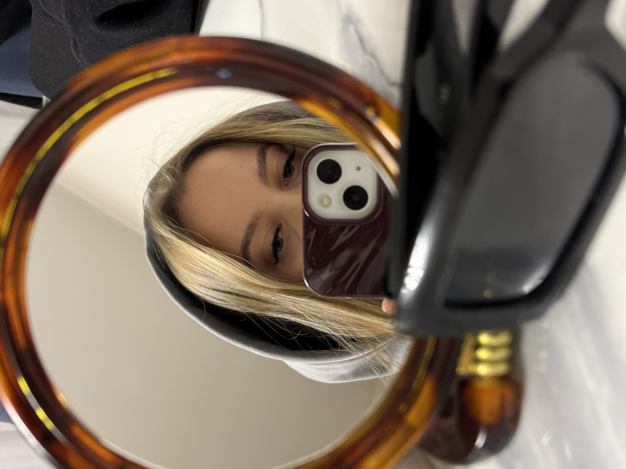The image is a rotated mirror selfie taken indoors, featuring a young person with blonde hair, visible in the reflection of a round mirror. The person is wearing a gray hooded sweatshirt, and their left eye, eyebrow, and prominent eyelashes are clearly seen. A smartphone with a dark red or burgundy case is held up, obscuring most of the person's face, with its dual-camera module and flash prominently reflected in the mirror.
The mirror has a distinct tortoiseshell-patterned frame in shades of amber and brown. To the right of the mirror, partially visible, are a pair of dark-framed sunglasses or eyeglasses, resting on a white surface that suggests a marble-like texture. The background reflected in the mirror is a plain, light-colored surface, possibly a wall. The activity depicted is the act of taking a self-portrait through a mirror. The setting is an indoor space, potentially a bathroom or vanity area, located in Krasnodar, Russia. No specific time of day or emotional expression is discernible, and there is no visible text in the image.