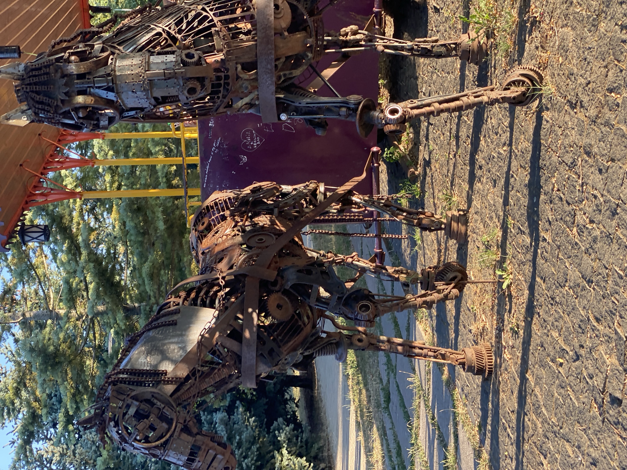The image shows two large, intricate sculptures of horses, constructed from what appears to be recycled metal parts, including gears, chains, and sheet metal. They are positioned outdoors on a paved surface that resembles cobblestones, with patches of grass and dirt in between. In the background, there are lush green pine trees and a structure with a reddish-orange roof supported by yellow poles, possibly part of an amusement ride or park attraction. The horse on the left is facing towards the left of the frame, while the horse on the right is facing towards the right. The sculptures exhibit a weathered, rusty patina, suggesting they have been exposed to the elements. The lighting indicates it is daytime, with sunlight casting shadows.
The specific location in Chișinău, Moldova, is not discernible from the image itself. However, the overall scene suggests an outdoor public space, possibly a park, a sculpture garden, or an entertainment venue. The horse sculptures are the primary subjects of the image, and their detailed construction from scrap metal is a key feature. There are no people visible in the frame. The environment is natural and somewhat rustic, with the metal sculptures providing a stark contrast to the surrounding greenery.