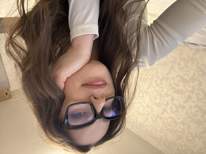 The image depicts a close-up portrait of a female individual in an indoor setting located in the city of Chaikovsky, Russia. The subject, appearing to be in her twenties or thirties, has long, wavy brown hair and is wearing black-framed glasses with a noticeable blue tint on the lenses. Her lips are a muted pink. She is dressed in a light-colored, likely off-white or light grey, long-sleeved ribbed top. Her left hand is positioned with curled fingers supporting her chin, while her gaze is directed upwards and to the right, accompanied by a slight smile. The background on the left features a wall covered in light beige wallpaper with an intricate, repeating damask or floral pattern. To the right, the background is darker and less defined, showing a light-colored, possibly grey, vertical surface and a warm light source emanating from the upper right corner. A light-colored rectangular object is faintly visible in the upper right quadrant of the background, and a soft, light-colored surface, possibly bedding, is partially visible on the far left.