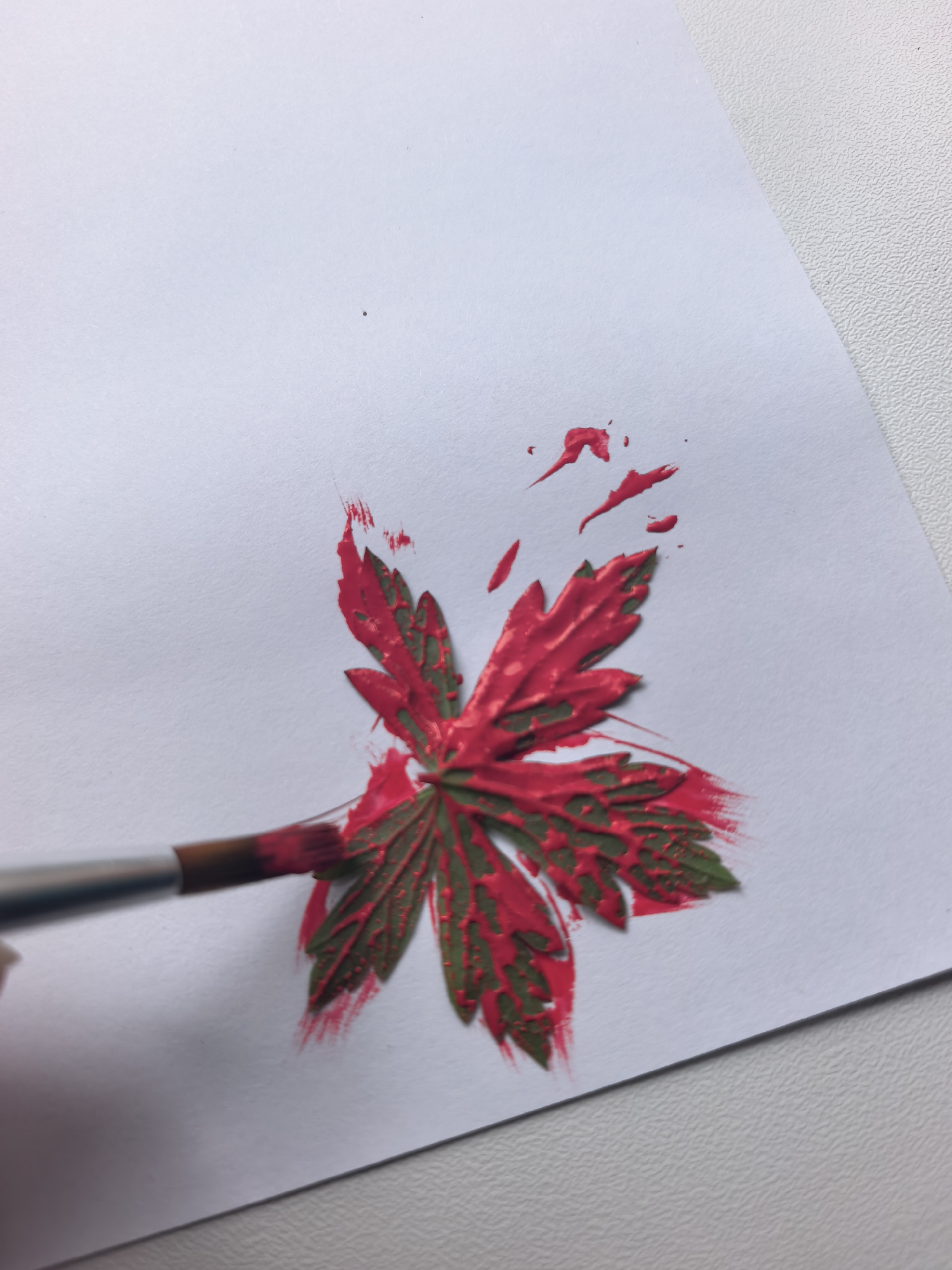 A close-up shot shows a paintbrush being used to apply red paint to a green leaf imprinted on white paper. The leaf is positioned in the lower center of the frame, with the paintbrush touching its lower left side. Red paint has splattered around the leaf and on the paper. The background is a textured white surface.