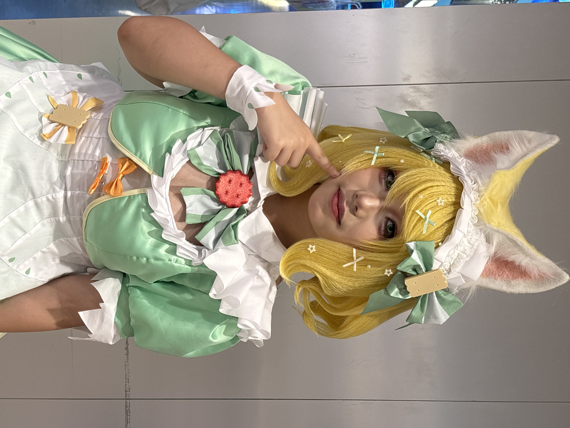 A person, angled vertically, is seen wearing a cosplay outfit. The cosplay includes a yellow wig with green bows and star accents, and a white headdress with yellow and pink bunny ears. The outfit is white and green, with a large red biscuit-like embellishment on the bodice. The person has green eyes. The background is a nondescript grey wall. The location is Unknown, Unknown. The person is pointing a finger at their cheek.