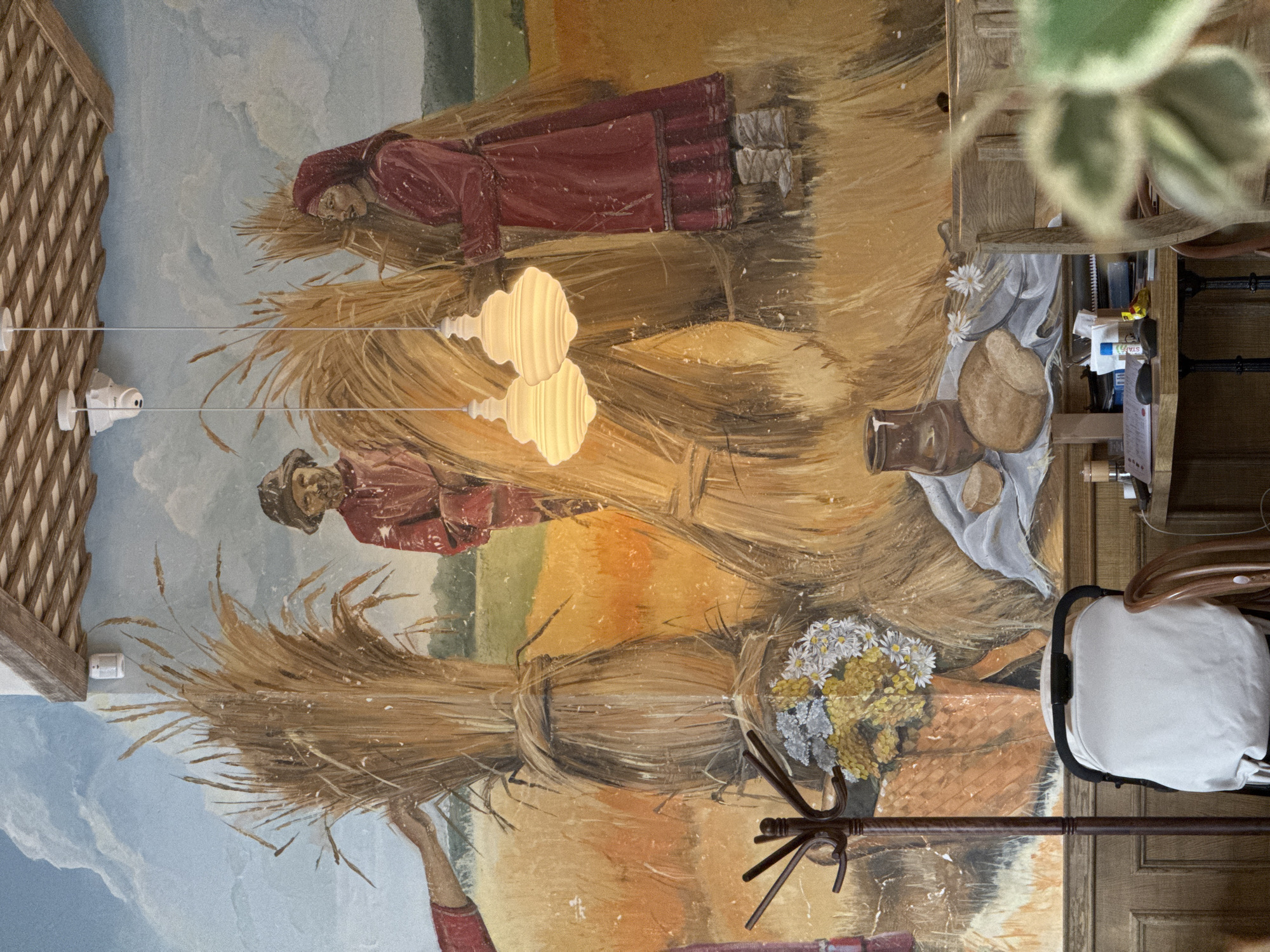 The image depicts a mural painting on a wall in what appears to be a restaurant or cafe. The location is presumably in the city of Izhevsk, Russia, though this is not explicitly confirmed in the image.
The mural depicts a scene reminiscent of peasant life, potentially inspired by Russian folklore or historical depictions of rural life. Two figures, likely women, are portrayed working or interacting with haystacks. They wear traditional clothing, including red garments. The haystacks are central to the composition.
In the foreground, a section of the mural shows a still life arrangement featuring a pitcher, bread, and wildflowers on a cloth. Other objects present include lighting fixtures, part of a wooden structure, and a chair.