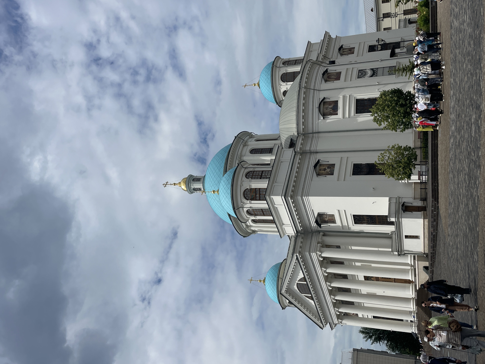 The image depicts the Annunciation Cathedral in Kazan, Russia, a white Orthodox church with blue domes and golden crosses. The cathedral features classical architecture with a prominent portico supported by columns, and several smaller domes topping its structure. In front of the cathedral, a paved plaza is occupied by a group of tourists, some of whom are gathered together, while others are walking. The sky above is partly cloudy, with a mix of gray and white clouds. The overall scene conveys a sense of religious and historical significance, with visitors exploring the architectural marvel.