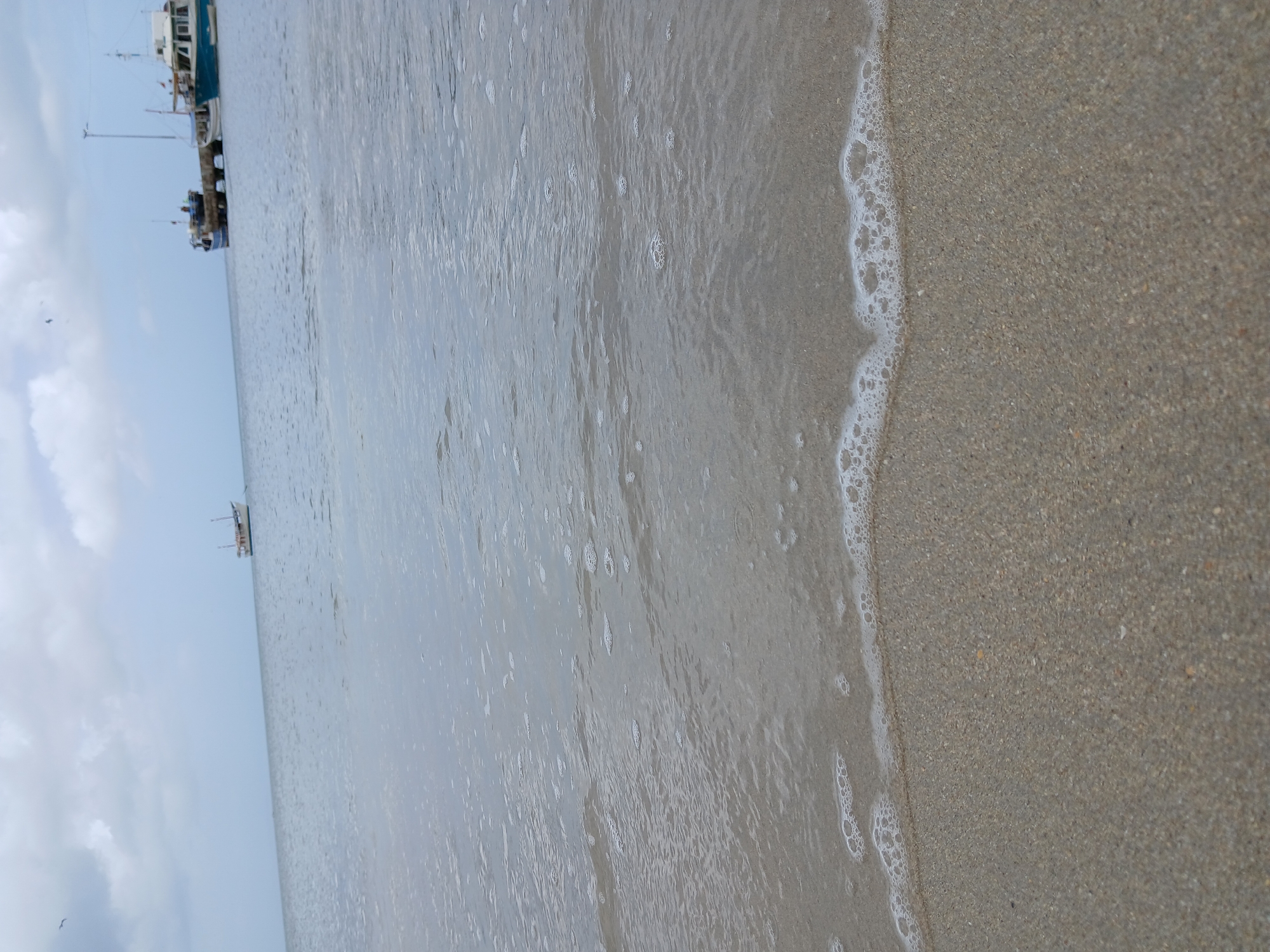 The image shows a beach scene in Juan Griego, Venezuela. The foreground features wet sand meeting the ocean's edge, with white foam from the waves visible. In the distance, under a cloudy sky, there are boats docked at a pier. The perspective is slightly tilted.