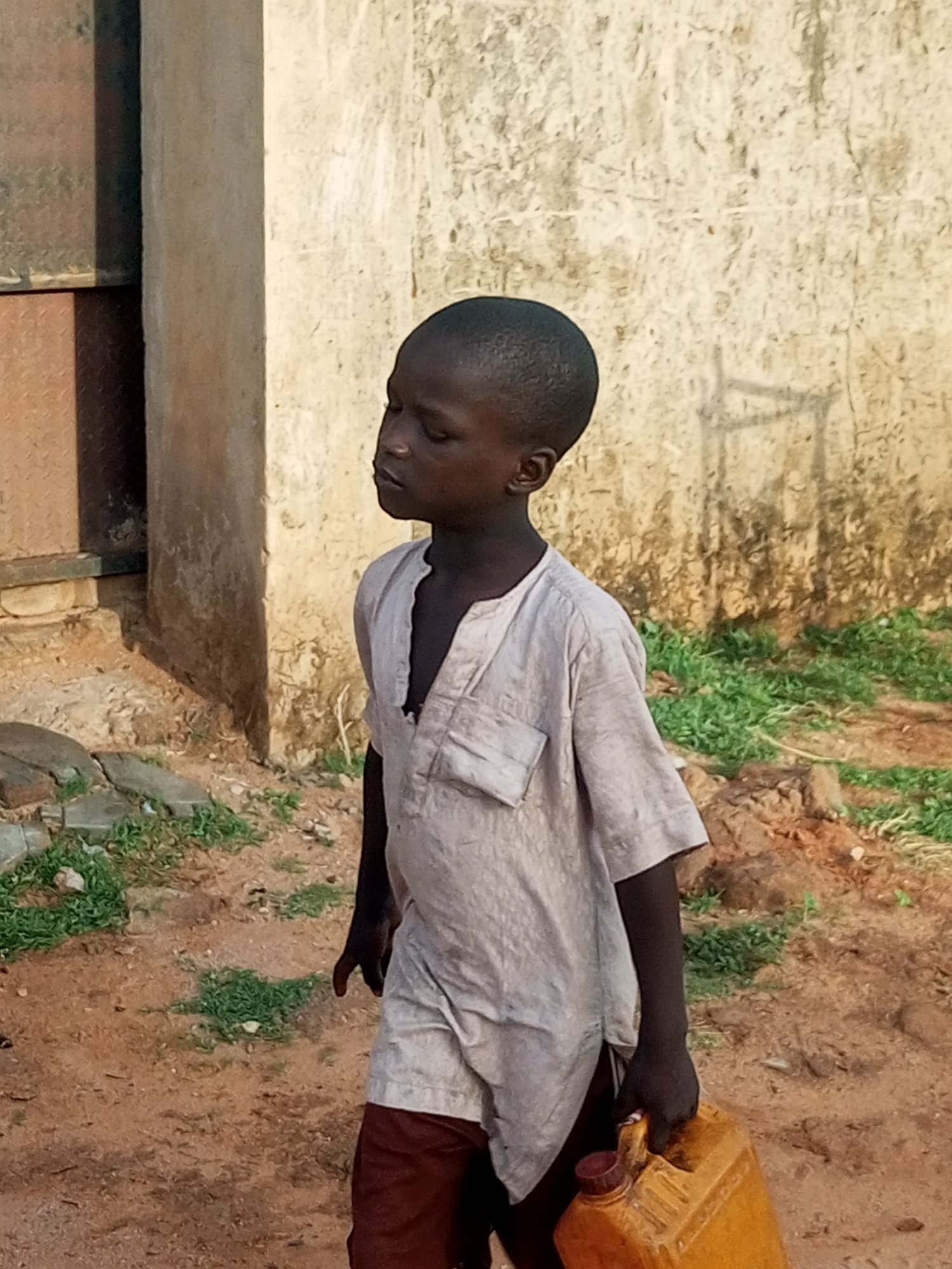 A young boy, approximately 5-10 years old, with dark skin and closely cropped dark hair, is depicted walking from right to left in an unknown location, likely a rural or semi-urban area. He is viewed from the chest up, slightly angled away from the camera, with his head turned to look towards his left. His eyes are slightly downcast, and his expression is neutral.
The boy wears a light-colored, possibly off-white or pale grey, loose-fitting tunic-style shirt with a V-neck and short sleeves. A patch pocket is visible on the left side of his chest. Below the tunic, dark reddish-brown trousers or shorts are visible. In his right hand, he carries a rectangular, yellow-orange plastic jerrycan-style container with a dark red or brown cap.
The ground is predominantly reddish-brown earth with sparse patches of green grass. In the background, to the left of the boy, a weathered building structure is visible, featuring a light-colored, possibly concrete or stucco wall alongside darker, textured panels that might be metal or a different building material. To the right of the boy, another light-colored, roughly textured wall, possibly stucco or weathered concrete, forms the background. The scene is illuminated by bright, natural daylight.