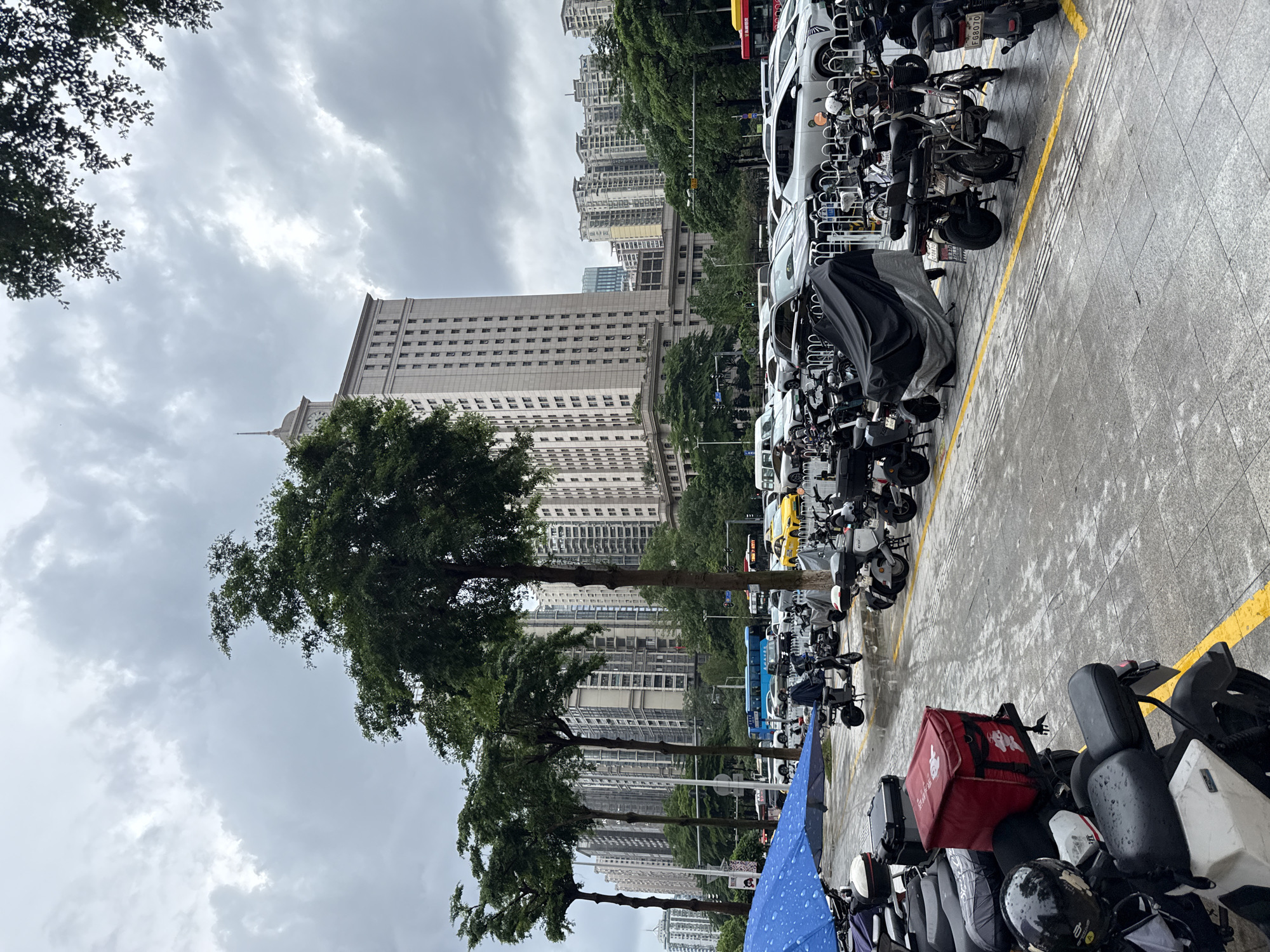 The image shows a street scene in Tianhe District, China. A row of parked electric scooters and motorcycles are visible in the foreground, some covered with tarps. A blue umbrella is open, suggesting it might be raining or has recently rained, as the ground appears wet. Behind the scooters, a street is lined with trees and various buildings, including a tall, light-colored skyscraper with a clock tower and other residential buildings. Cars are also parked and driving on the street. The sky is overcast and cloudy.