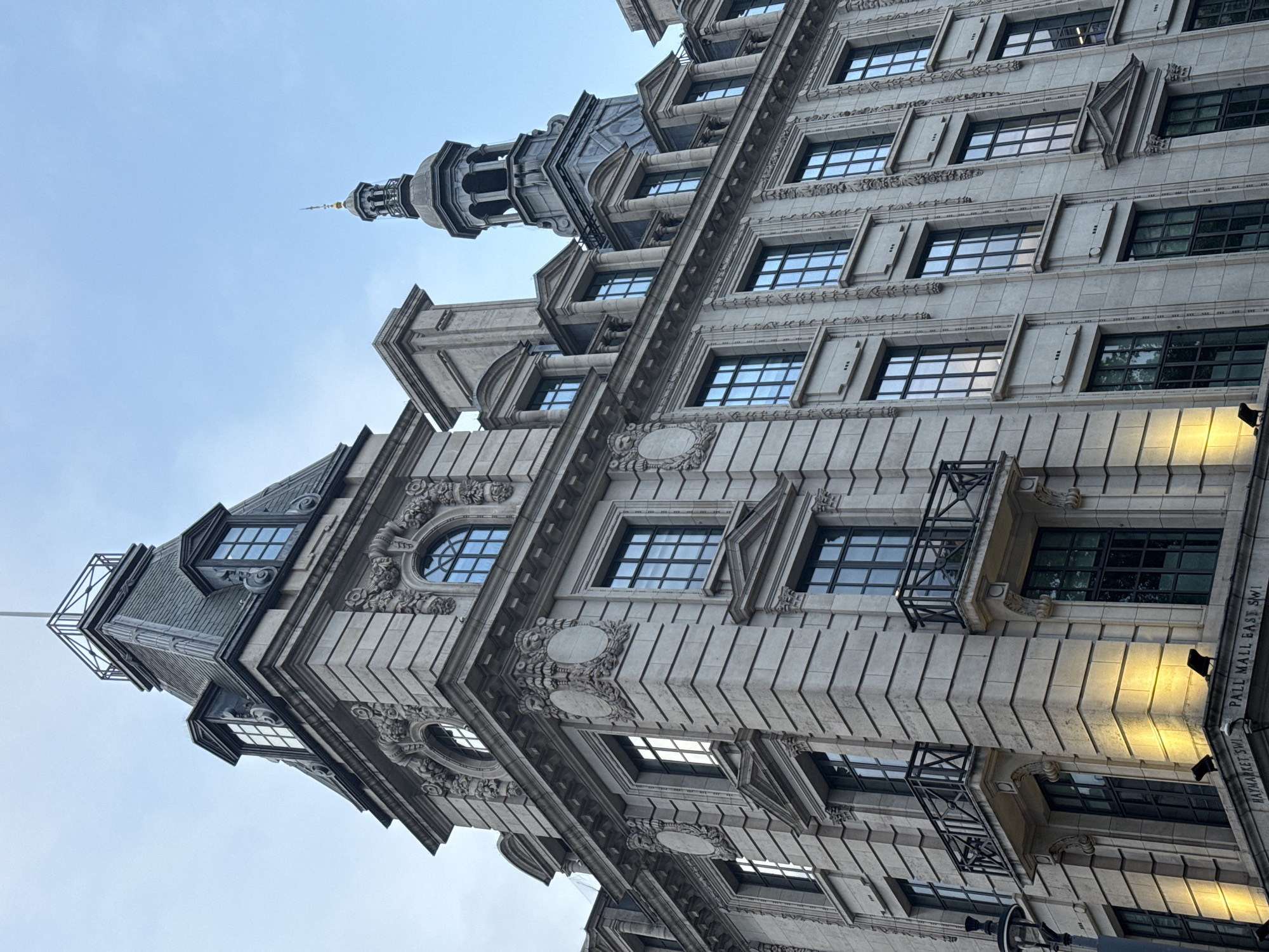 The image is a low-angle shot of a grand, ornate building located in London, United Kingdom. The building, presumably constructed of stone, features a complex facade with multiple windows, decorative carvings, and projecting architectural elements. A tower-like structure rises from the corner of the building, topped with a spire. The facade details include pilasters, ornamental roundels, and horizontal bands of textured masonry. Some windows have small balconies. The words "PALL MALL EAST SW1" and "HAYMARKET SW1" are partially visible on the building's facade, indicating its location near Pall Mall and Haymarket in central London. The sky above is overcast, with a light blue hue. The perspective of the shot emphasizes the building's height and grandeur.