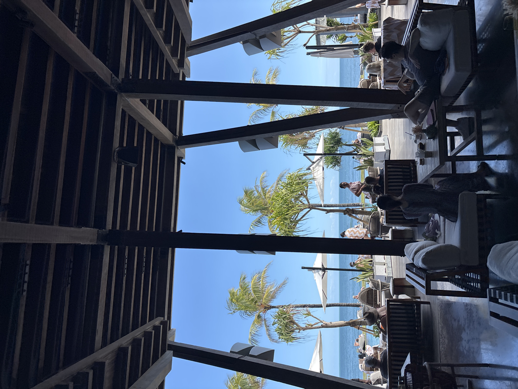The image captures a view from under the wooden structure of a restaurant or resort, looking out towards the beach and ocean in North Kuta, Indonesia. The wooden beams create a framed perspective of the scene. Outside the structure, the clear blue sky meets the light blue ocean. Palm trees sway in the breeze along the shoreline. People are seen lounging on outdoor furniture, including chairs and sofas. Some are sitting at tables, enjoying drinks. There are also white parasols scattered across the beach area, providing shade.