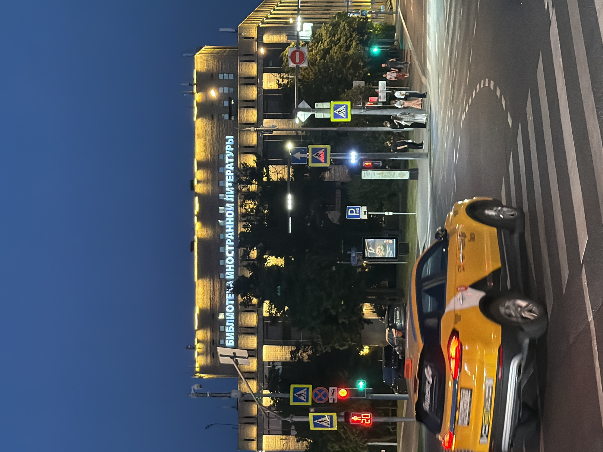 The image captures a street scene in Moscow, Russia, at dusk. A yellow taxi is crossing a pedestrian crossing in the foreground. Behind the taxi, a multi-story building with illuminated windows and a large neon sign displaying Cyrillic text is visible. The sign reads "БИБЛИОТЕКА ИНОСТРАННОЙ ЛИТЕРАТУРЫ," which translates to "FOREIGN LITERATURE LIBRARY." Traffic lights, pedestrian crossing signs, and streetlights are present, indicating an urban intersection. Several people are standing on the sidewalk near the crosswalk, and trees line the street. The sky is a deep blue, characteristic of twilight.