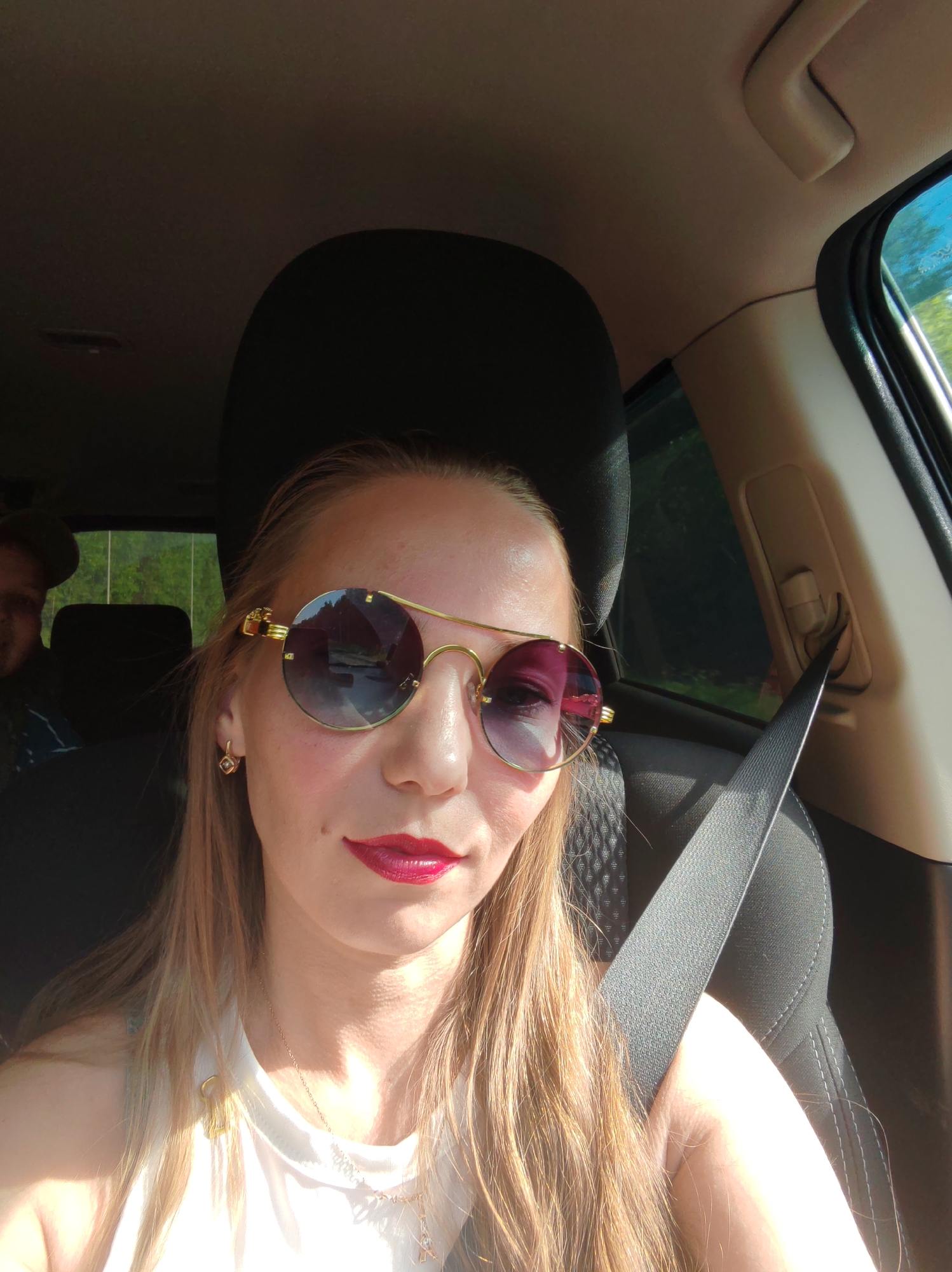 A woman with blonde hair, wearing round sunglasses and red lipstick, is taking a selfie in the passenger seat of a car. A man wearing a cap is visible in the background, in the driver's seat. The car interior is visible, including a seatbelt. The windows show a green, wooded landscape. The image was taken on a sunny day, with light reflecting off the woman's hair and the car's interior. The location is identified as Vylygort, Russia.