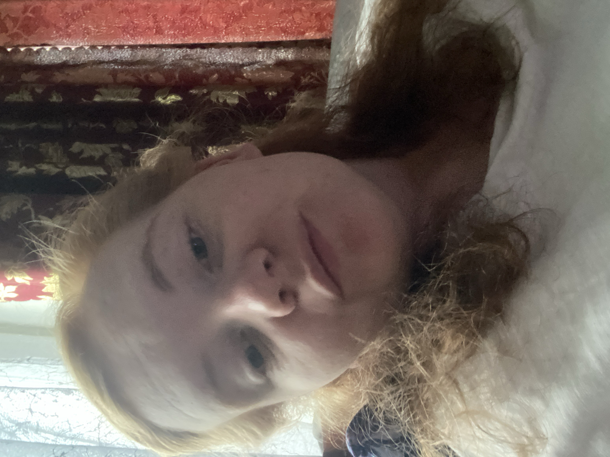 A close-up portrait features a young individual, likely female, with fair skin and light-colored, possibly curly or wavy, hair. The person is positioned with their head at the top of the frame, lying down and looking directly into the camera with a neutral to slightly relaxed expression, lips slightly parted.
In the background, behind the person's head, is a distinct red fabric, possibly a curtain or piece of furniture, embellished with a repeating gold, ornate pattern. Below this, a darker, patterned area suggests further decorative elements. To the left of the person, a soft, white or light-colored fabric, likely a pillow or bedding, partially frames their face and hair. The setting appears to be indoors, possibly a bedroom or a cozy living space, given the intimate angle and soft fabrics. The lighting is gentle and even, highlighting the person's features without harsh shadows. No specific activity beyond posing for the portrait is evident. There is no visible text in the image. While the provided location context is Vyatskiye Polyany, Russia, the image itself contains no explicit geographical markers.