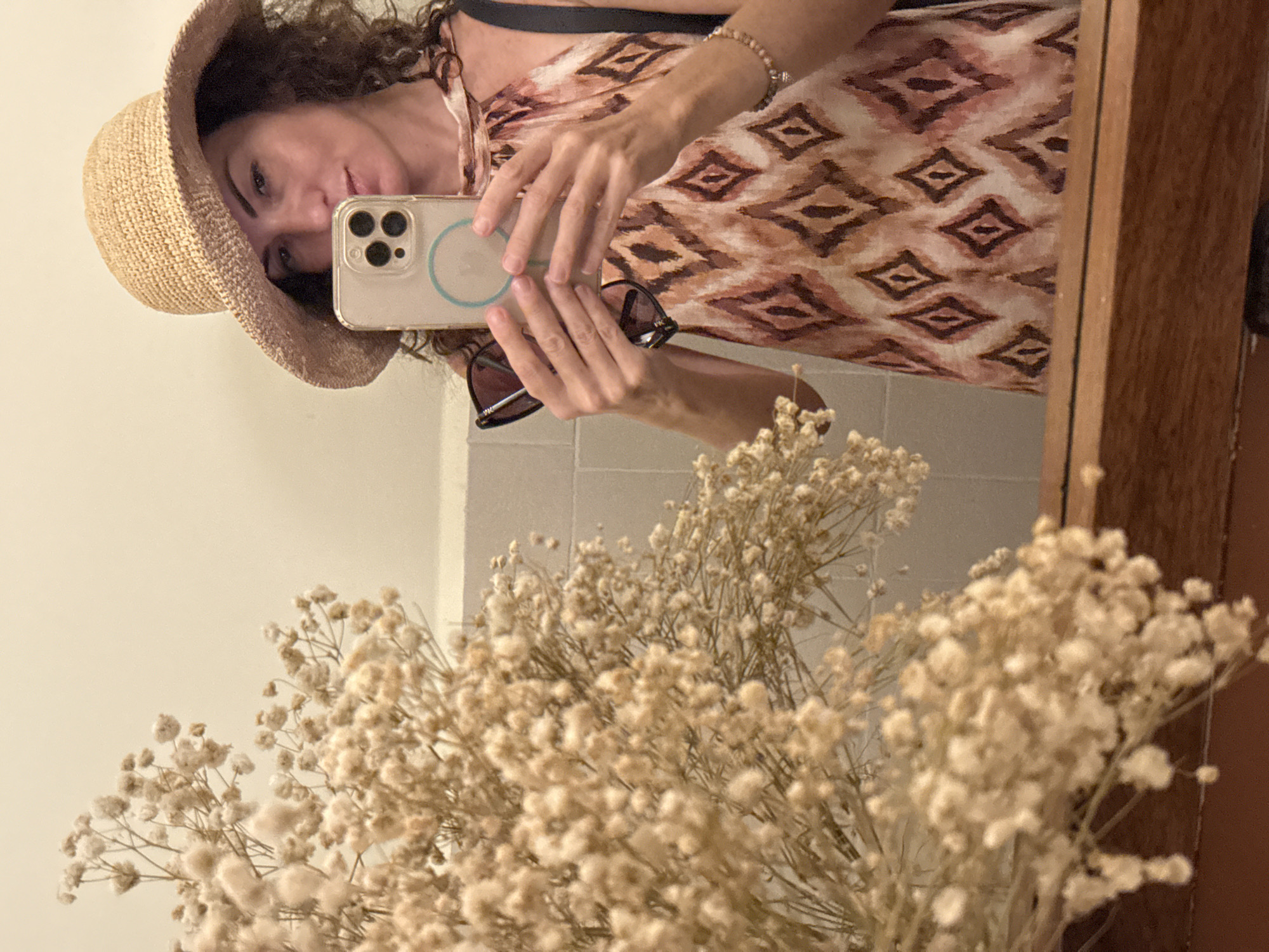 A woman wearing a straw hat and a patterned dress takes a mirror selfie in a room. She holds a phone in her right hand and a pair of sunglasses in her left. Dried flowers are visible in the foreground. The background appears to be a bathroom with tiled walls. The reflection shows the woman's face and upper body. The image is taken in Nha Trang, Vietnam.