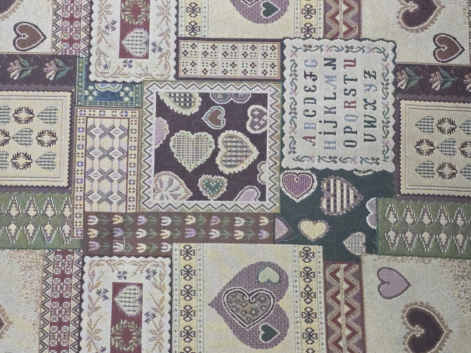 The image is a close-up, overhead shot of a tapestry or fabric with a quilted patchwork design. The design features various embroidered elements, including stylized hearts, floral motifs, geometric patterns, and an alphabet sampler. The colors are muted, with shades of beige, brown, green, purple, and red. There are no people, animals, or recognizable landmarks in the image. It is impossible to determine the location in Ulan-Ude, Russia, based on this image.