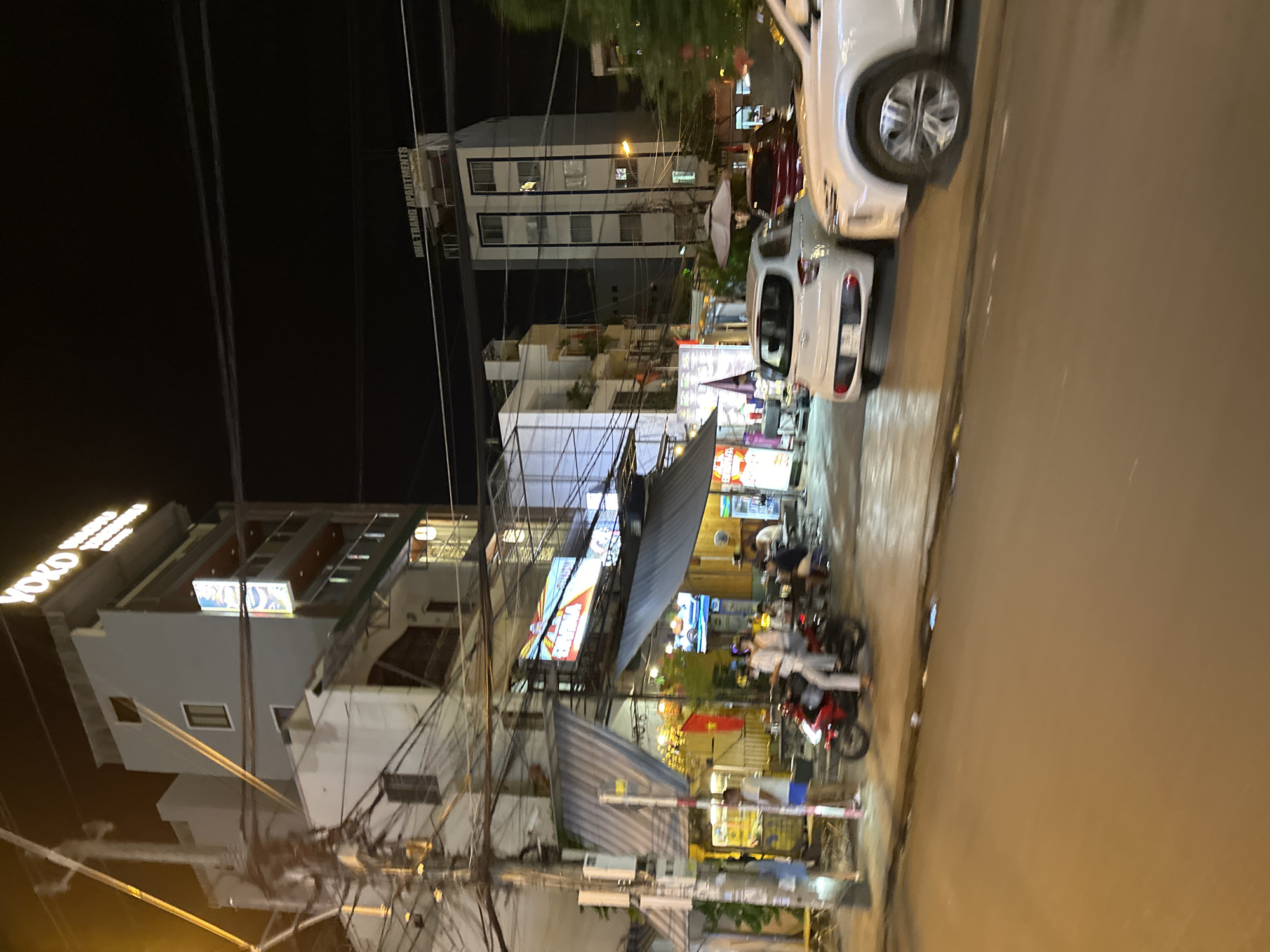 The image captures a nighttime street scene in Nha Trang, Vietnam. A tall building with a "YOKO HOTEL" sign is visible on the left. Adjacent to it, a row of businesses with awnings and outdoor seating lines the street. Several people are seated at tables, and a person is riding a red motorcycle. Two white cars are parked on the right side of the street, with a third car partially visible behind them. Another building, identified as "NHA TRANG APARTMENTS" by a sign, stands further down the street on the right. The street is paved, and utility poles with numerous wires are prominent on the left side of the image. The lighting suggests it is evening or night, with artificial lights illuminating the businesses and buildings.