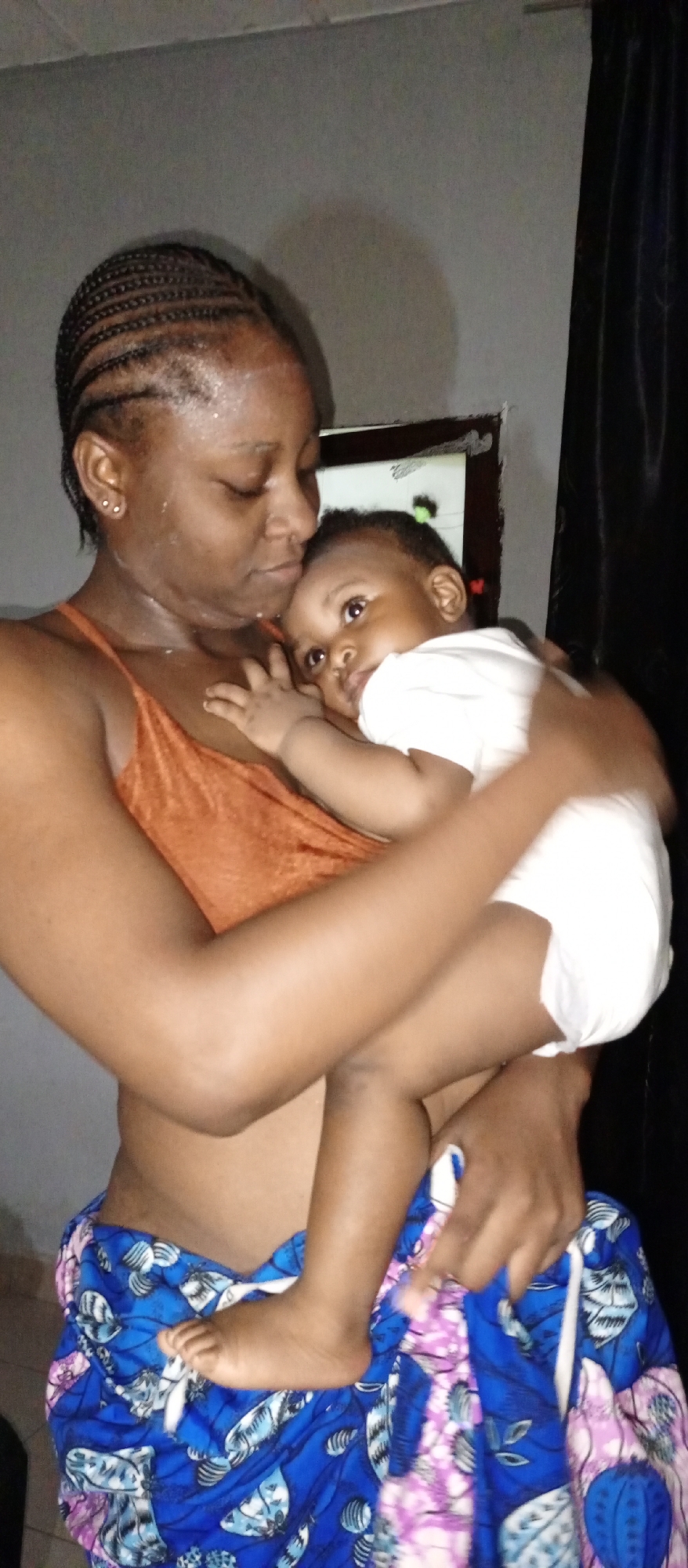 The image depicts a woman holding a baby. The woman has dark skin, braided hair, and is wearing an orange sleeveless top and a blue and purple patterned wrapper around her waist. She is looking down at the baby. The baby is fair-skinned and wearing a white outfit. The baby is looking towards the camera. The background appears to be indoors, with a gray wall, a window and dark curtains. The location is presumed to be in Keffi, Nigeria.