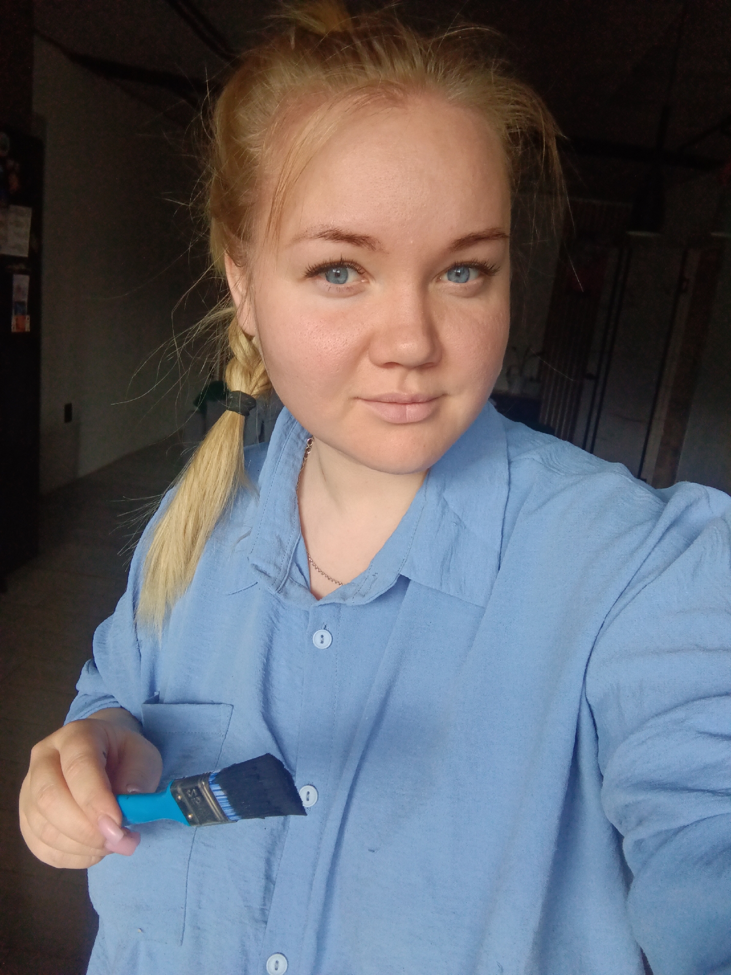 The image is a portrait of a young woman, likely in Фоки, Russia, featuring her upper body and head. She has blonde hair, pulled back into what appears to be a messy bun with a braid secured by a black hair tie visible on her left side. Her bright blue eyes are directed at the camera, and she maintains a neutral, calm expression. She is dressed in a light blue, long-sleeved button-up shirt, and a thin gold chain necklace is visible around her neck.
In her right hand, she holds a paint brush with a bright blue handle and dark bristles, suggesting it has been used with dark paint or is new with dark bristles. Text "BRUSH" and "1.5" are clearly visible on the brush handle, and "GOLDEN A" can be partially seen on the metal ferrule of the brush.
The background is dark and appears to be an indoor, utilitarian space such as a garage, workshop, or storage room, indicated by indistinct dark objects like a cabinet or refrigerator on the left and shelving or storage racks on the right. The overall lighting is somewhat dim, which highlights the woman in the foreground. No active event is depicted beyond her holding the paintbrush and posing for the photograph.