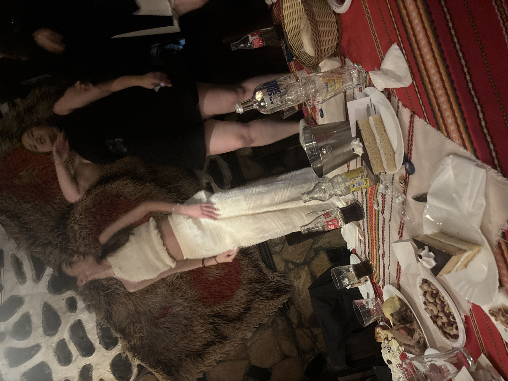 This image depicts an indoor social gathering, likely in a traditional restaurant setting, in Blagoevgrad, Bulgaria. The background features a stone wall, partially obscured by a large, shaggy brown fur-like wall hanging with a prominent red patch on its left side.
Two women are prominently positioned in the upper half of the frame. The woman on the left, with dark hair, wears a light-colored, possibly off-white, halter-neck crop top and matching wide-leg trousers. She is looking to her right (viewer's left) with her right arm slightly raised. To her right, a second woman with dark hair wears a black sleeveless dress and a dark belt. Her right arm is raised, and her left hand holds a small, light-colored object. She is looking upwards with her mouth slightly open. Additional figures are partially visible in the dimly lit background.
A table in the lower half of the image is covered with a red and white striped tablecloth adorned with horizontal patterns in various colors. The table is laden with food and beverages. Visible items include an empty bottle of Absolut Vodka, two glass Coca-Cola bottles (one partially full/empty, one full), several clear drinking glasses (some containing a dark liquid), and a silver ice bucket. Food items present are a plate with roasted meat and mashed potatoes, a platter of mixed nuts (cashews, almonds, and peanuts), multiple slices of layered cake with cream and chocolate topping served on white paper plates, and a wicker basket containing bread rolls. White paper napkins and a blue bottle opener are also on the table. The overall lighting is dim, creating a warm, ambient atmosphere.