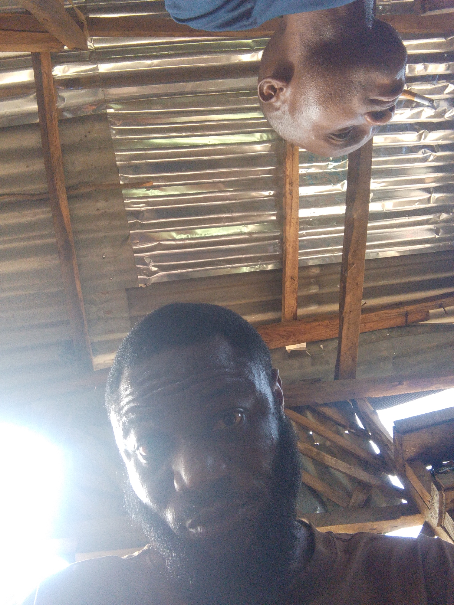 The image shows two men under a corrugated metal roof supported by wooden beams. One man, with a shaved head, is upside down and has a lit cigarette in his mouth. The other man, with a beard, is facing the camera. The location is likely in Lokoja, Nigeria, given the prompt's specification. The light conditions suggest an outdoor environment with sunlight filtering through gaps in the roofing structure.