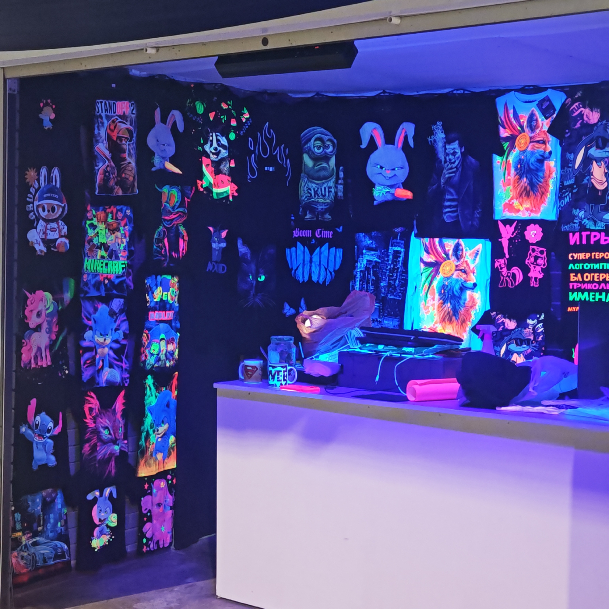 The image captures an indoor retail space, likely a shop or stall, featuring a vibrant display of merchandise under ultraviolet (blacklight) illumination. Numerous black t-shirts are hung on a dark wall, each showcasing designs that glow brightly in neon colors.
The displayed designs include a variety of popular culture references and themes:
- **Cartoon/Game Characters:** Stitch, multiple bunny characters (some holding carrots), a Minion labeled "SKUF", a My Little Pony unicorn, Sonic the Hedgehog, a fox with elaborate headwear, and a character resembling The Joker. Designs related to "STANDOFF 2", "MINECRAFT", and "ROBLOX" are also visible.
- **Other Graphics:** A raccoon holding watermelon slices, a cat with glowing green eyes, cityscapes, a sports car, flames, and abstract patterns. One shirt has "Boom Time" text.
- **Counter:** A white counter or table is positioned in the foreground, illuminated by the UV light, giving it a purple hue. On the counter, there's a clear jar, a mug with a Superman logo, and another mug with the text "VELO". A pink rolled mat and some plastic bags are also visible.
A sign on the right side of the display features Russian text: "ИГРЬ" (Games), "СУПЕР ГЕРО" (Superheroes), "Логотип" (Logos), "БЛОГЕРЬ" (Bloggers), "ПРИКОЛЬ" (Cool/Funny), and "ИМЕНА" (Names), with "MYA" also present.
The blacklight creates a striking contrast, making the fluorescent designs pop against the dark background, giving the scene a dynamic and engaging atmosphere. The setting appears to be a commercial establishment in городское поселение Щелкино, Ukraine.