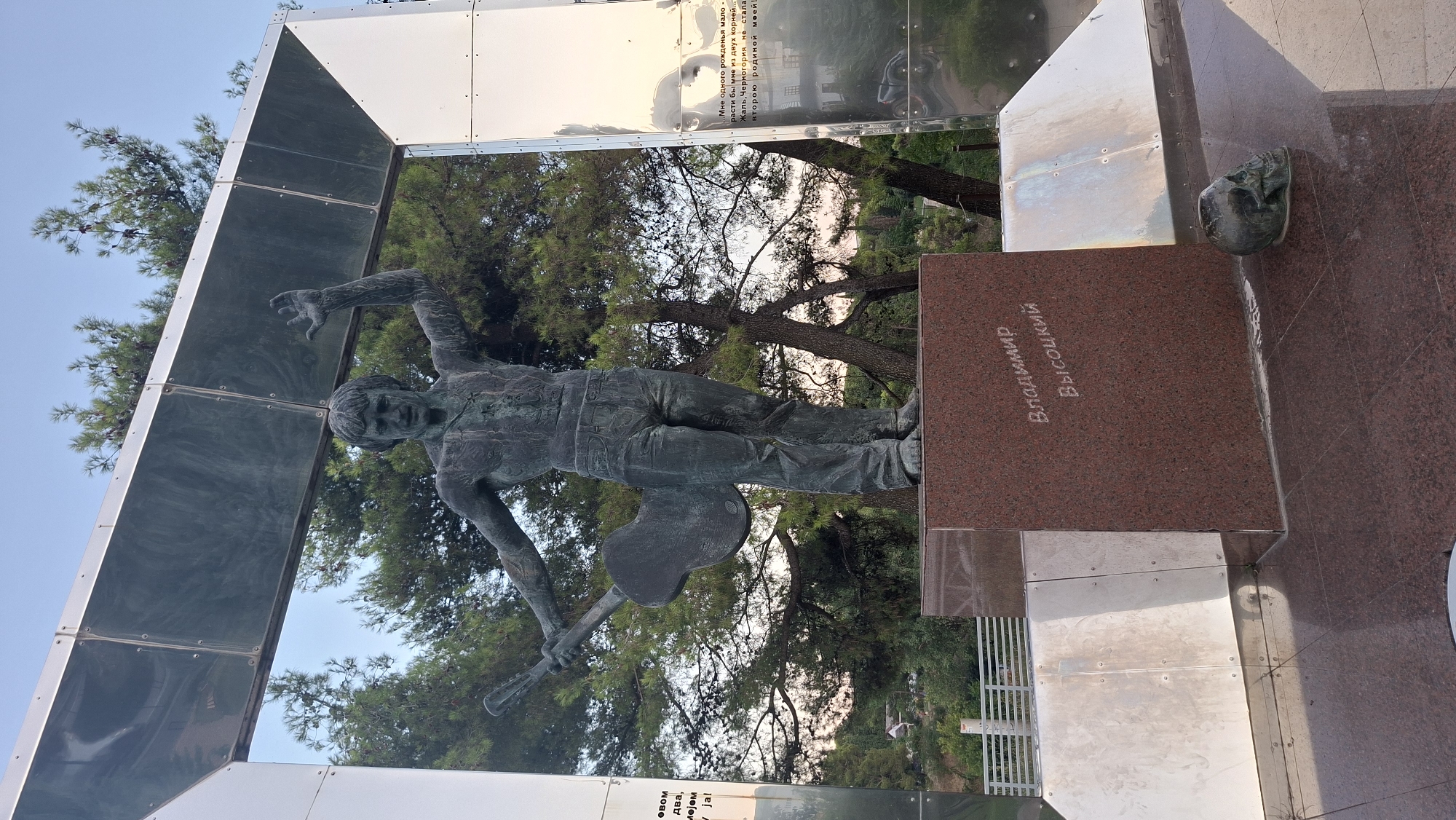 The image depicts a monument to Vladimir Vysotsky in Podgorica, Montenegro. A bronze statue of Vysotsky, holding a guitar, stands on a red granite pedestal inscribed with his name in Cyrillic. The monument is framed by a metallic structure with reflective surfaces. A dark, skull-shaped object rests on the ground in front of the pedestal. Trees and a clear sky are visible in the background.