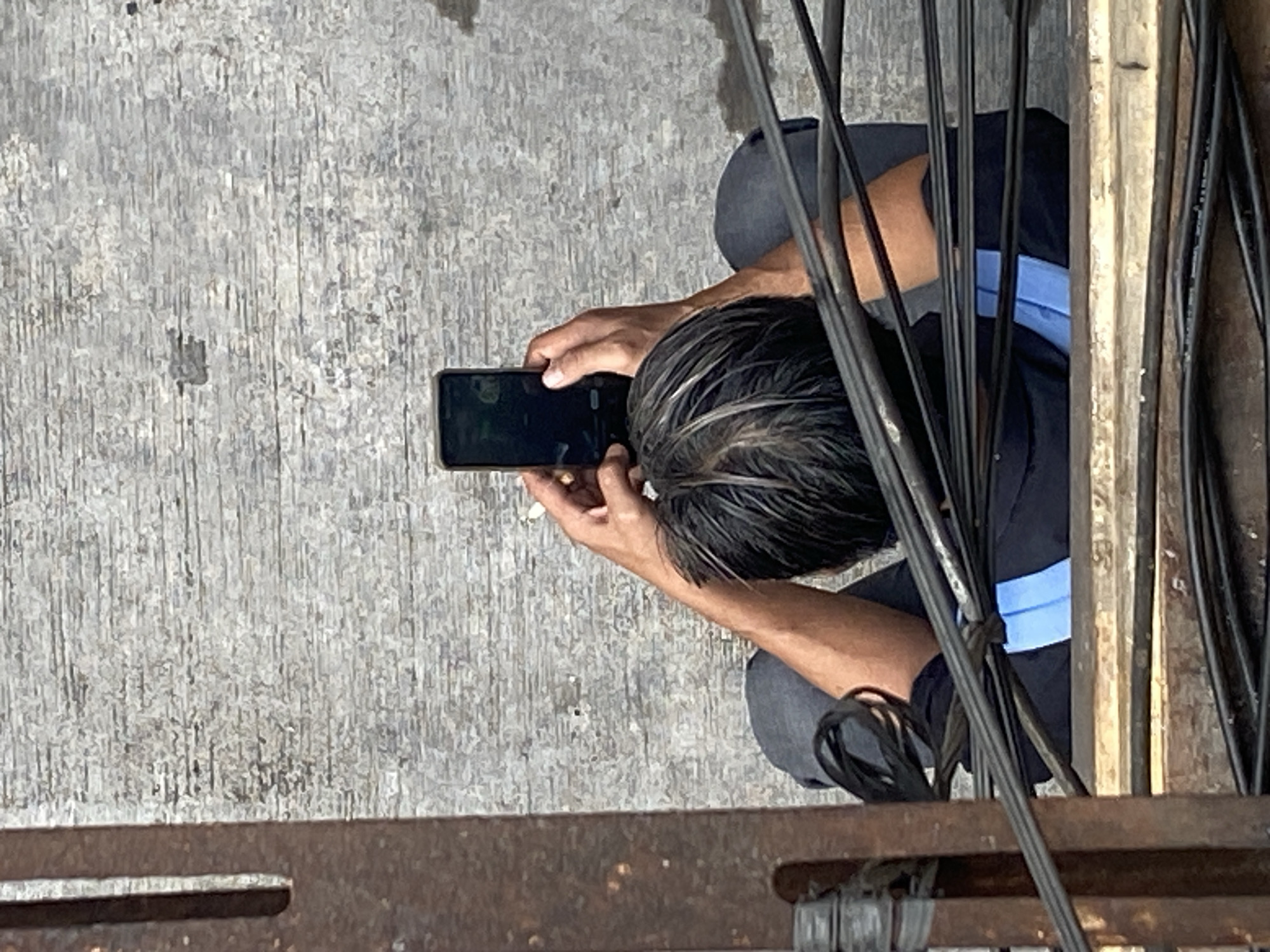 A person is viewed from above, sitting and holding a smartphone in portrait orientation, seemingly taking a picture or recording a video. The individual's back is to the camera, with their head tilted down towards the phone. They are wearing a dark shirt with a light blue accent on the collar area. The person's arms are visible, bent at the elbows as they hold the device. The background is a textured, gray surface, possibly concrete or a similar material, showing horizontal striations. In the foreground and to the left of the image, a metal structure with a rusted appearance is visible, along with a bundle of dark electrical wires. The scene appears to be outdoors, with the lighting suggesting daylight. The presence of electrical wires and the overall setting could indicate an urban or industrial environment within North Jakarta, Indonesia.