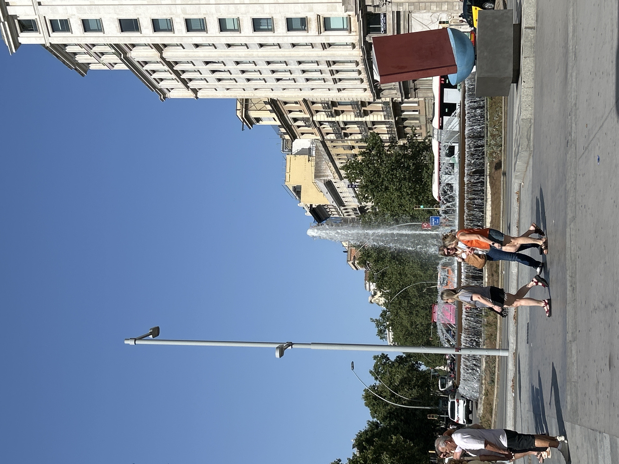 This is a photograph of a street scene in Barcelona, Spain, on a sunny day with a clear blue sky. In the foreground, there are several people walking on a paved area. To the left, an older man wearing a light-colored t-shirt and dark shorts is standing. Further to the right, three women are walking. One woman in a grey t-shirt and black skirt is walking away from the camera. Two other women, one in an orange tank top and blue shorts, and another in a white top and blue jeans, are walking towards the right side of the frame.
In the midground, a large, circular fountain with multiple jets of water is visible. Behind the fountain, there are trees and buildings lining the street. A tall street lamp stands to the left of the fountain.
In the background, a large, multi-story building with many windows and ornate architectural details dominates the right side of the frame. To the left of this building, other buildings with balconies and awnings are visible, suggesting a dense urban environment. A red, abstract sculpture resembling a sail on a boat is positioned in front of the building on the right.
Vehicles, including cars and a bus, are visible on the street behind the fountain. The overall impression is a busy urban square with pedestrian activity and public amenities.