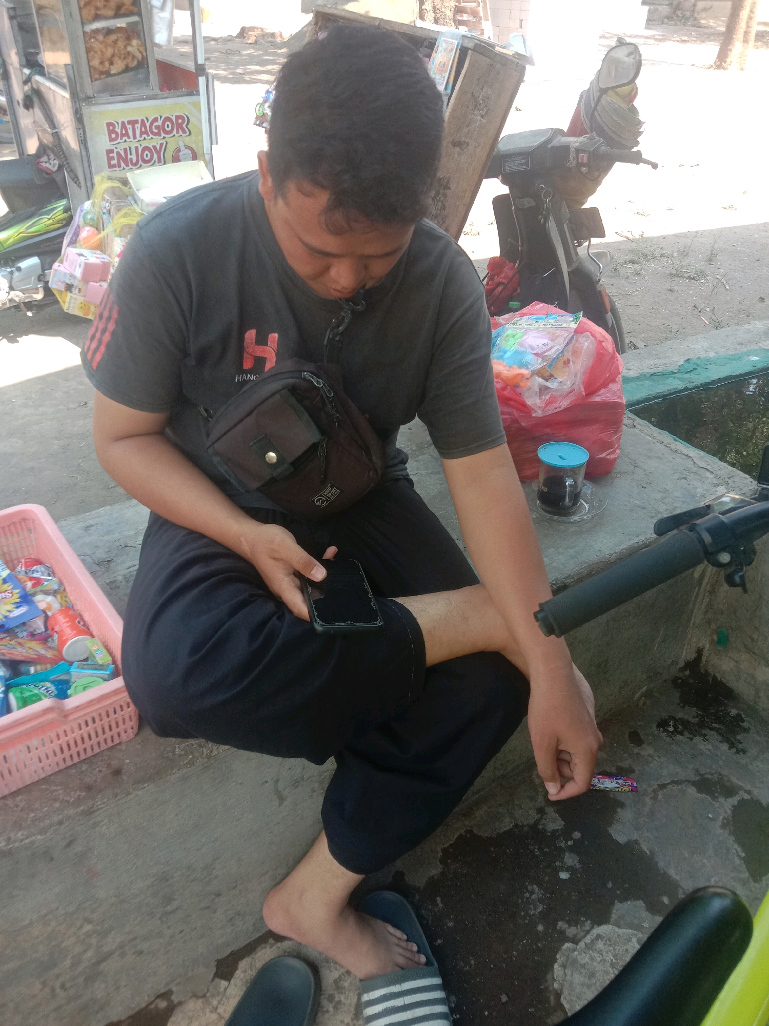 A man is sitting on a concrete step in what appears to be a street or roadside area, possibly in Ngempit, Indonesia. He is wearing a dark t-shirt and dark pants, and a small dark bag is slung across his chest. He is looking down at a smartphone he is holding in his right hand. To his left, a pink plastic basket is filled with various small packages of snacks and drinks. Behind him, there is a food cart with items on display, including a sign that reads "BATAGOR ENJOY." Further behind the man and to his right, a dark-colored motorcycle is parked. On the concrete ledge near the man, there is a clear glass with a blue lid containing a dark liquid, likely coffee. The ground is wet in places, suggesting recent rain. The overall lighting indicates it is daytime and sunny.