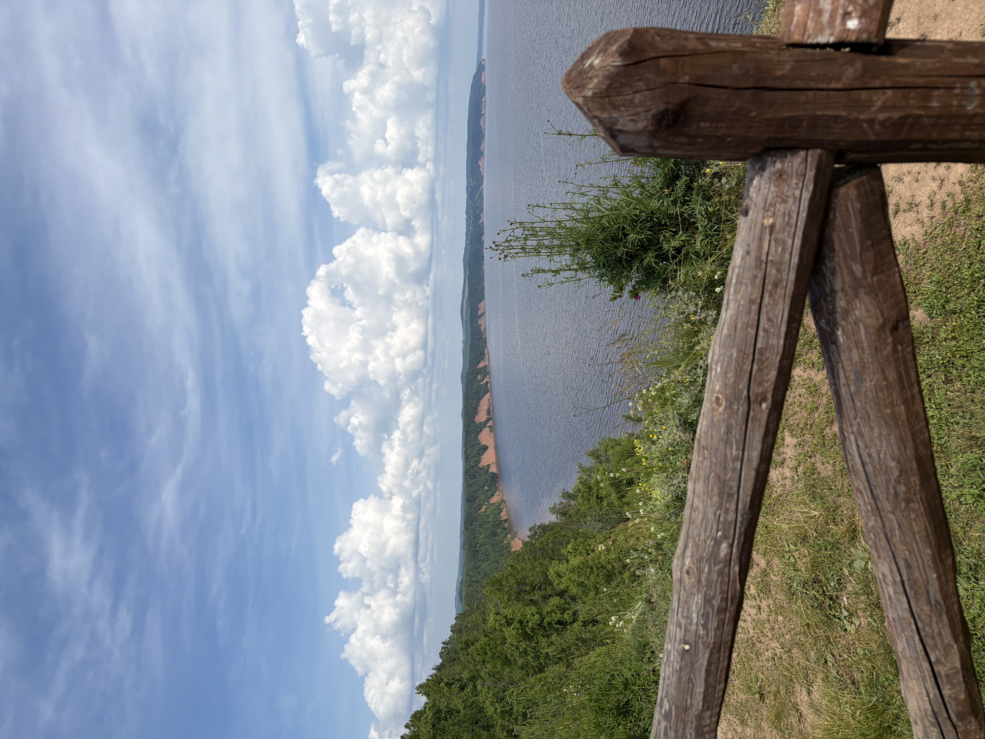 This image captures a panoramic view of a wide river flowing through a lush, green landscape under a partly cloudy sky. In the foreground, a rustic wooden fence made of rough-hewn logs runs horizontally across the frame, partially obscuring the view. Beyond the fence, a grassy slope descends towards the water, dotted with small wildflowers and vegetation. The river itself is a deep, calm blue, stretching towards a distant, tree-lined horizon. The far bank of the river features a prominent, reddish-brown cliff face interspersed with greenery. The sky is a vibrant blue, adorned with fluffy white cumulus clouds and wisps of cirrus. The overall impression is one of natural beauty and serenity, likely taken from an elevated viewpoint overlooking the river. The location in the city of Костоваты, Russia is not directly depicted or inferable from the visual elements within the image.