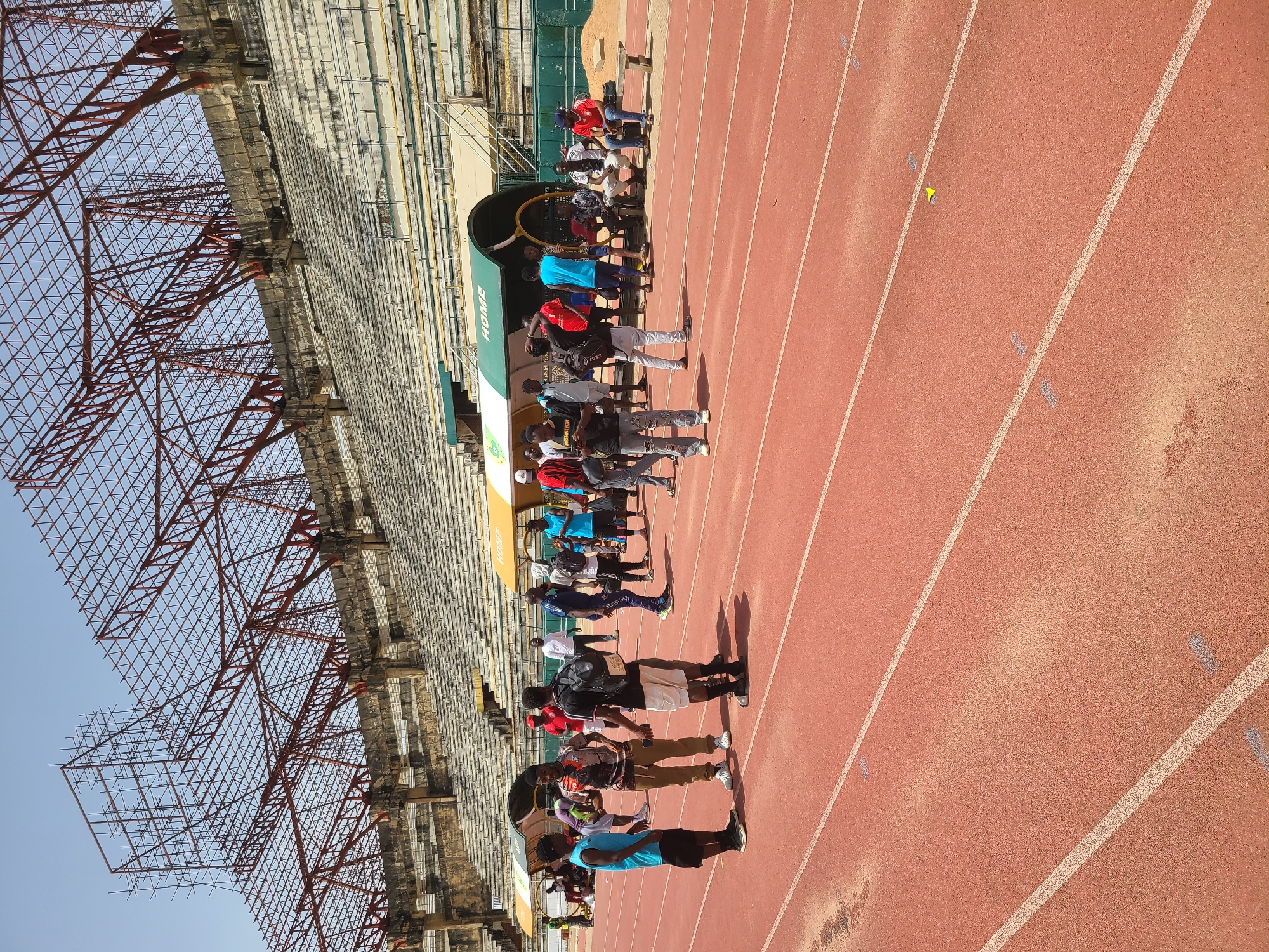 The image shows a group of people standing along a track at the New Stadium in Nigeria. The track is a reddish-brown color and has white lane markers. There are people standing on the track near a covered dugout with "HOME" printed on the side. A stadium structure with scaffolding is visible in the background. The sky is blue.