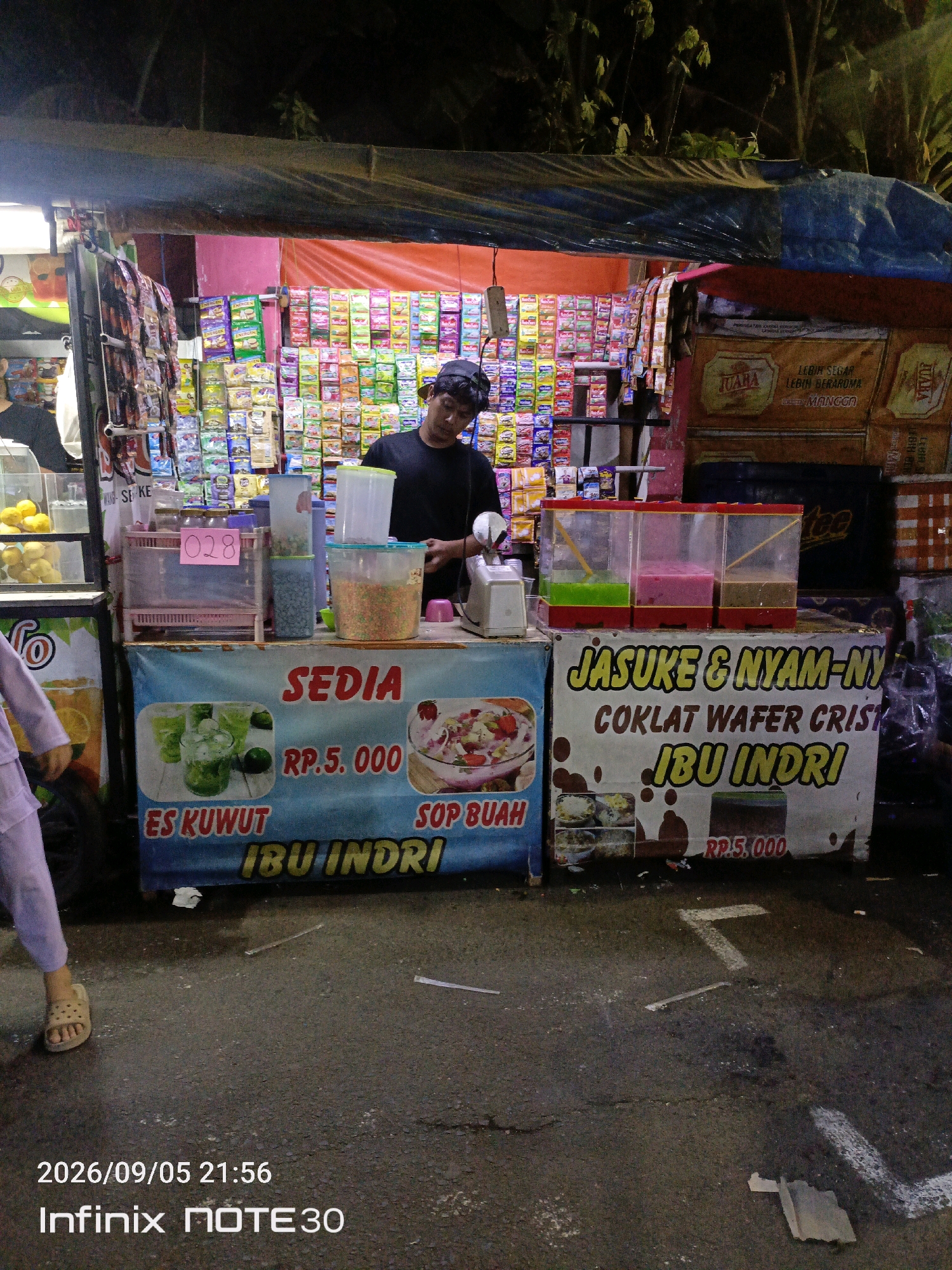 The image shows a street food stall at night in East Jakarta, Indonesia. The stall is selling various drinks and snacks. In the foreground, a person wearing light purple clothing and sandals is walking away from the camera. The stall itself is brightly lit, with shelves stocked with packaged goods and containers of colorful drinks. A man in a black shirt and hat is standing behind the counter, serving a customer. Signs on the stall advertise "ES KUWUT," "SOP BUAH," and "JASUKE & NYAM-NY," along with prices. The ground is asphalt, with some debris and markings visible. The overall impression is that of a busy night market.