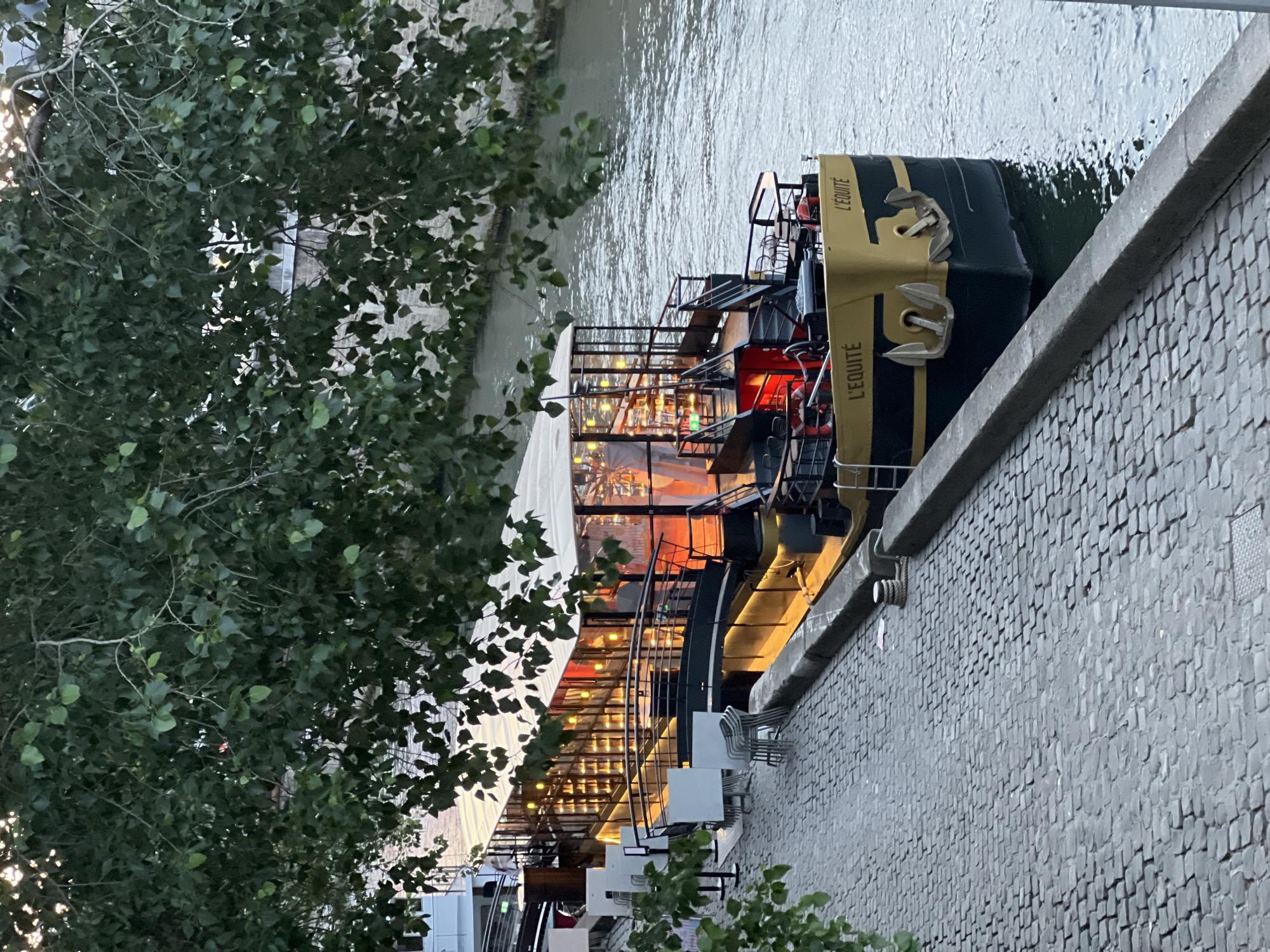 The image presents an elevated view of a boat, identified as "L'EQUITE," moored alongside a stone embankment on a river at dusk or early evening. Lush, dark green tree foliage heavily frames the left side of the scene, partially obscuring the river and the boat.
The boat itself features a dark hull with distinctive gold trim and prominent anchor designs on its side. Its interior is brightly lit with warm, golden lights, visible through large glass panels, suggesting it functions as a restaurant, bar, or event space. Adjacent to the boat, on the textured grey stone embankment, several white outdoor tables and chairs are arranged, indicating an al fresco seating area. The river's surface appears calm with subtle ripples, reflecting the ambient light.
No people are visible in the frame, contributing to a tranquil atmosphere. The visible text "L'EQUITE" is displayed prominently on the boat's exterior in gold lettering. The setting, with its characteristic river and stone embankment, is consistent with riverside scenes found in Paris, France. The warm artificial lighting against the diminishing daylight marks the time as evening.